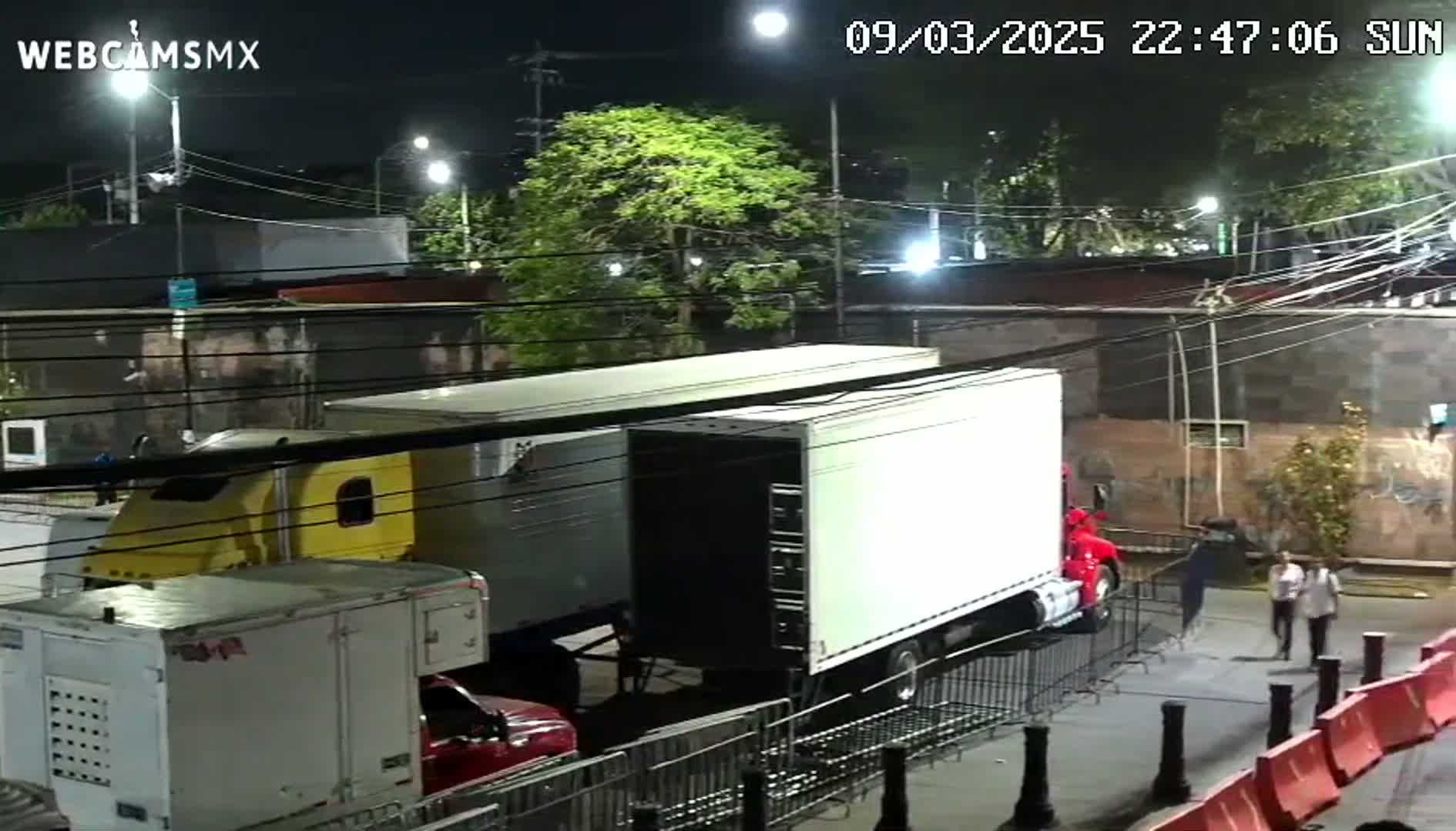 Several large trailers are parked at night on a city street, with a few pedestrians walking nearby under a moonlit sky.