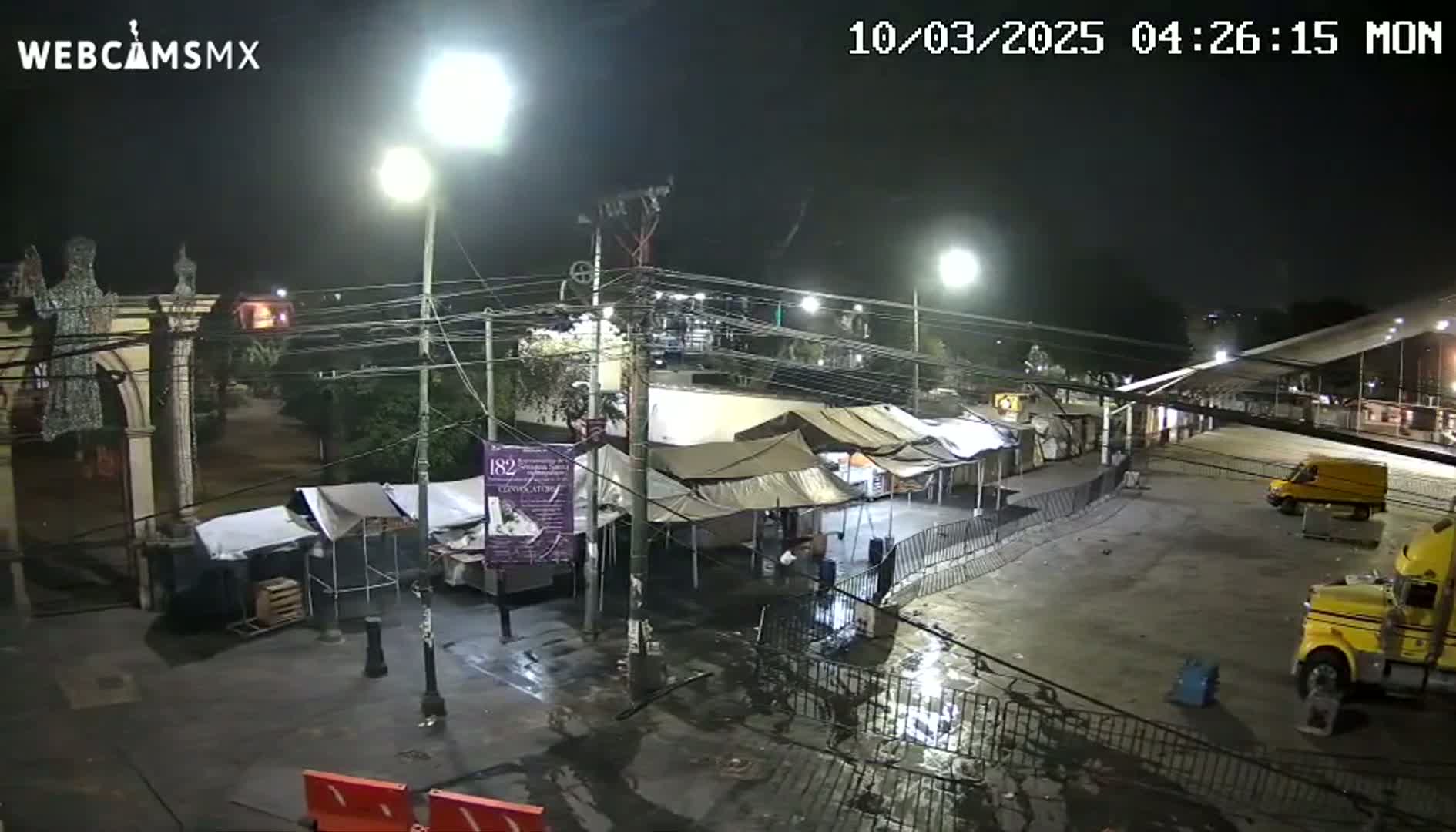 A nighttime, wet street scene shows temporary tents and stalls set up in front of a stone archway with statues, along with a few vehicles, including a large yellow semi-truck.