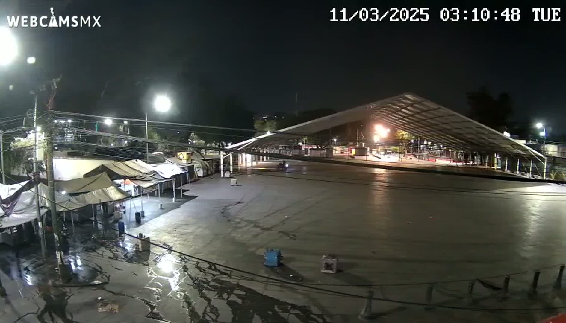 A large, empty paved area at night, illuminated by artificial lights, is partially covered by a large metal structure; a few tents and puddles are visible in the foreground.