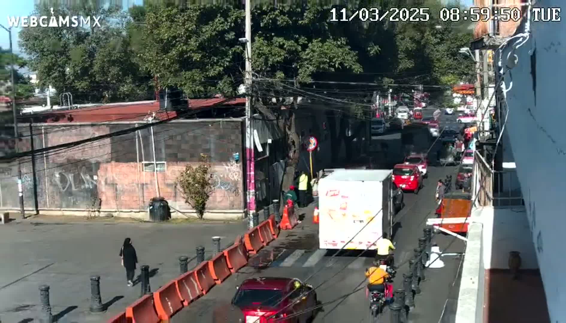 A street scene on a sunny day shows a busy road with cars and bicycles, people walking on the sidewalk, and construction barriers along a section of the street.