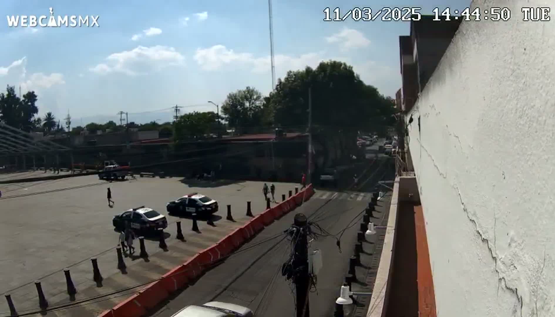 A high-angle view of a city street scene under a partly cloudy sky shows several parked police cars in a parking lot, pedestrians walking, and a few cars on the road.