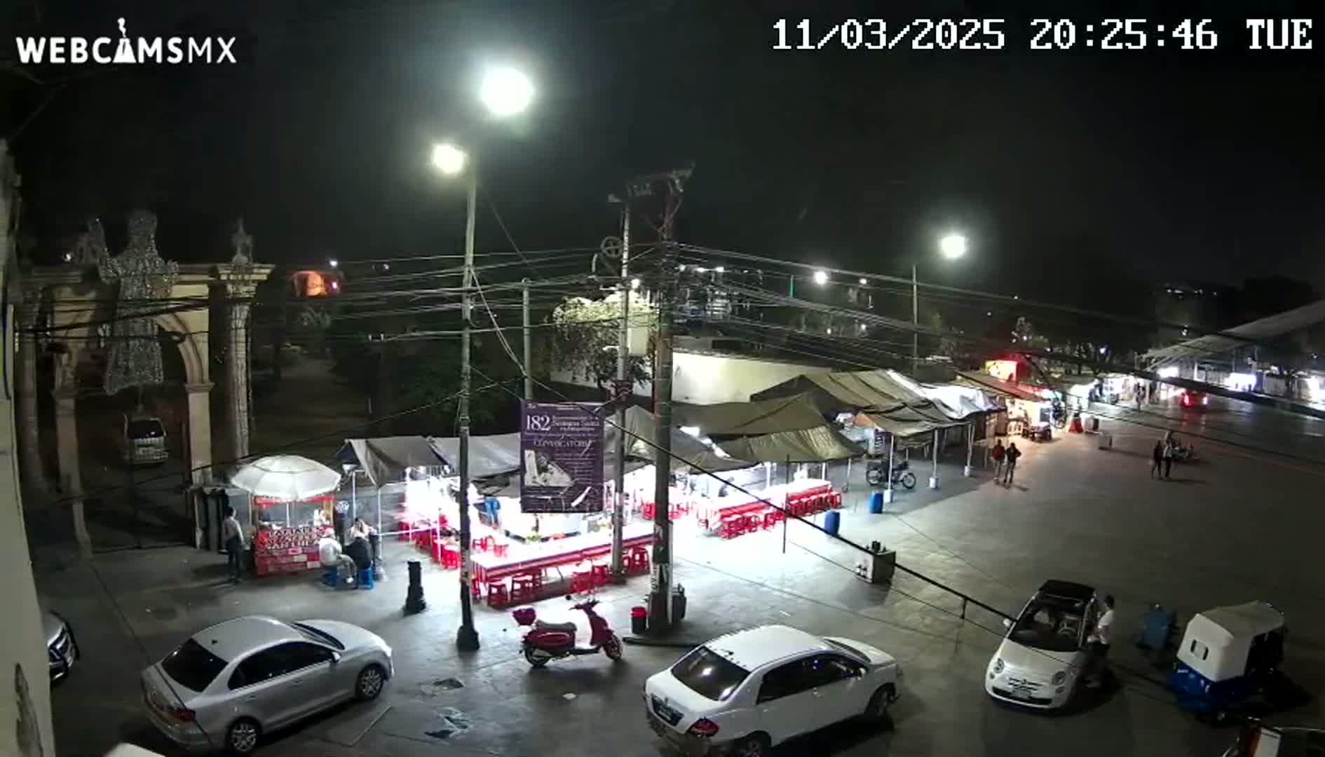 A nighttime aerial view of an outdoor market with numerous food stalls, cars, and people under clear skies.