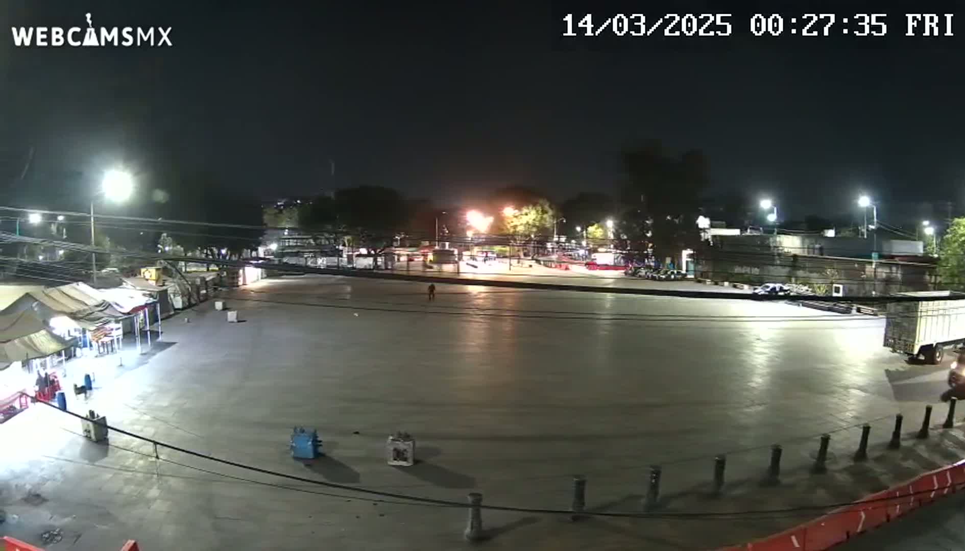 A nighttime, brightly lit, mostly empty plaza with a few vehicles, some temporary structures, and a lone pedestrian.