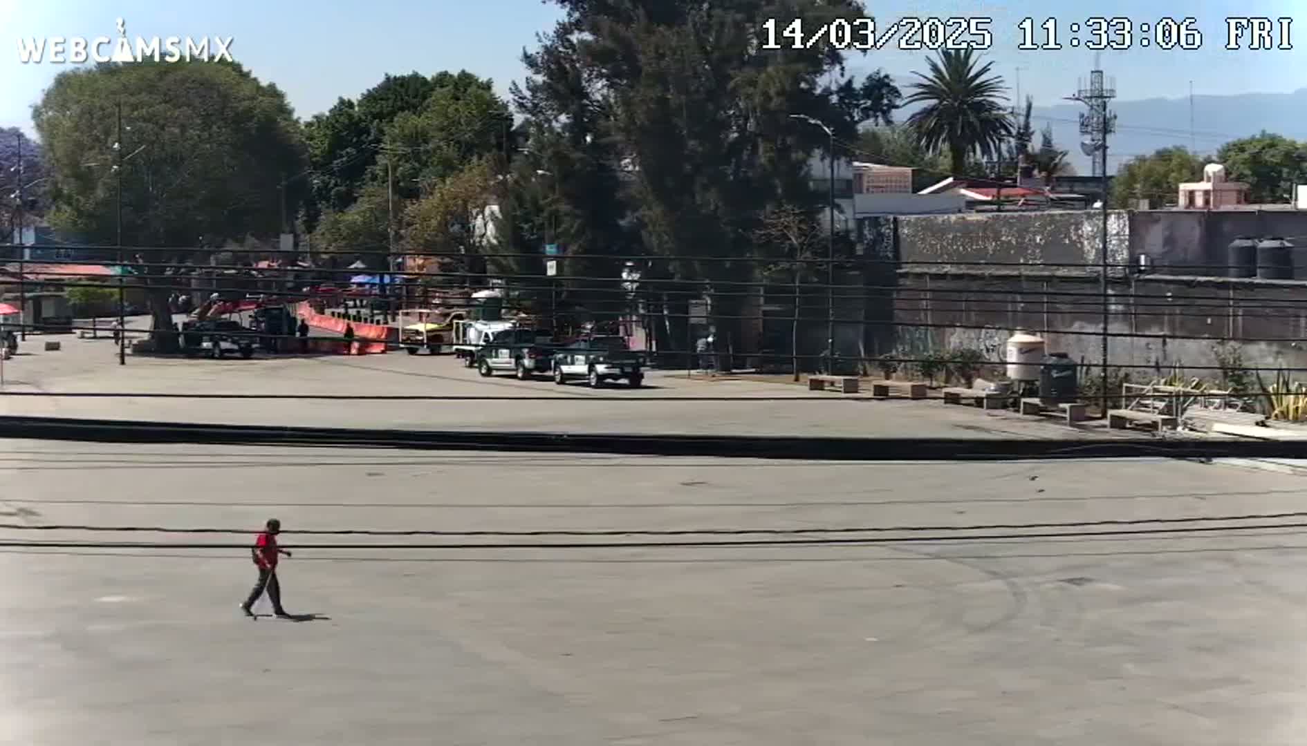 A man walks across a paved area in front of a building and several parked vehicles under a sunny sky.