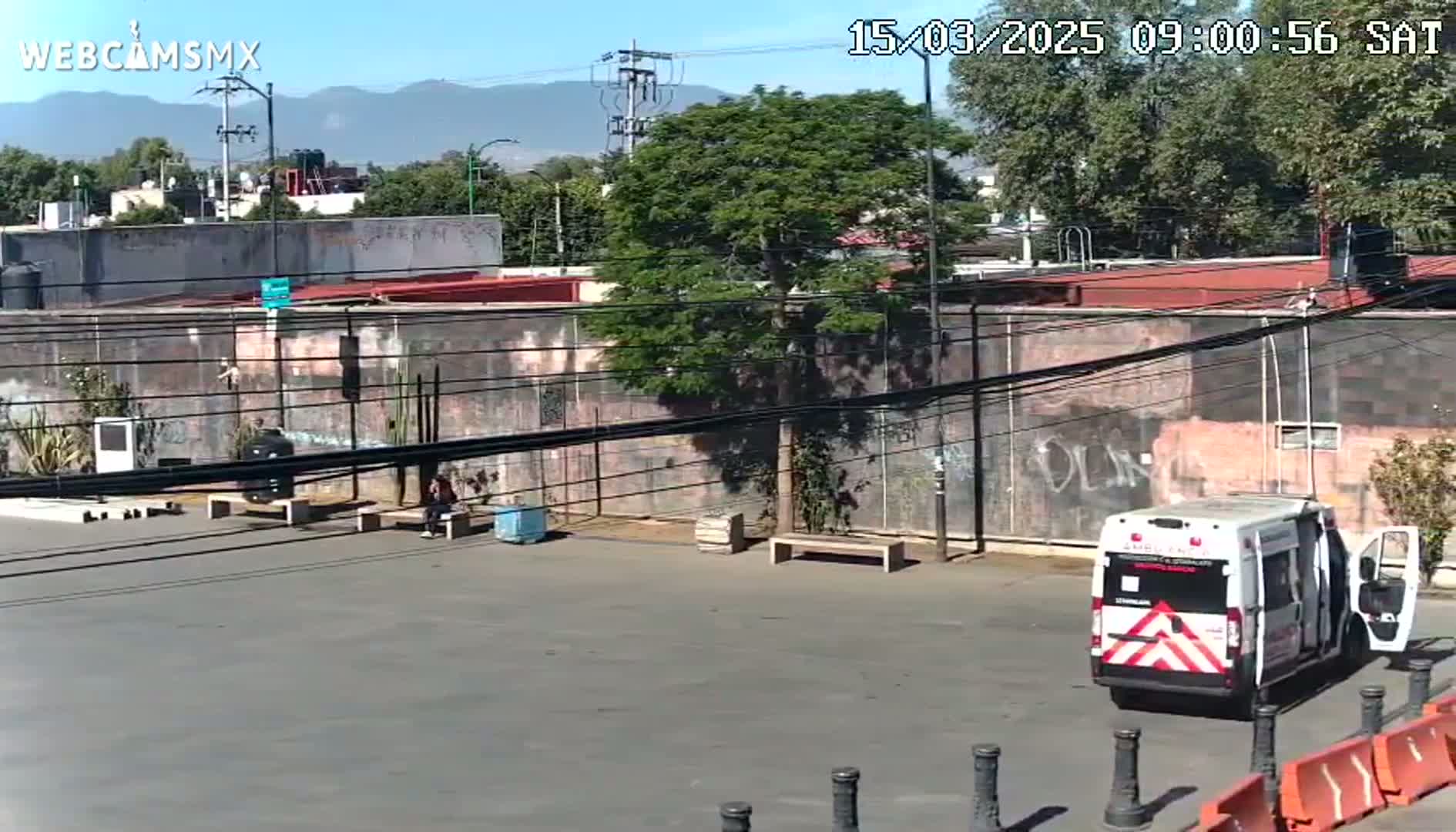 A mostly sunny day shows a paved area with a few people, some buildings with graffiti, a tree, and a medical van parked near orange and black barriers.