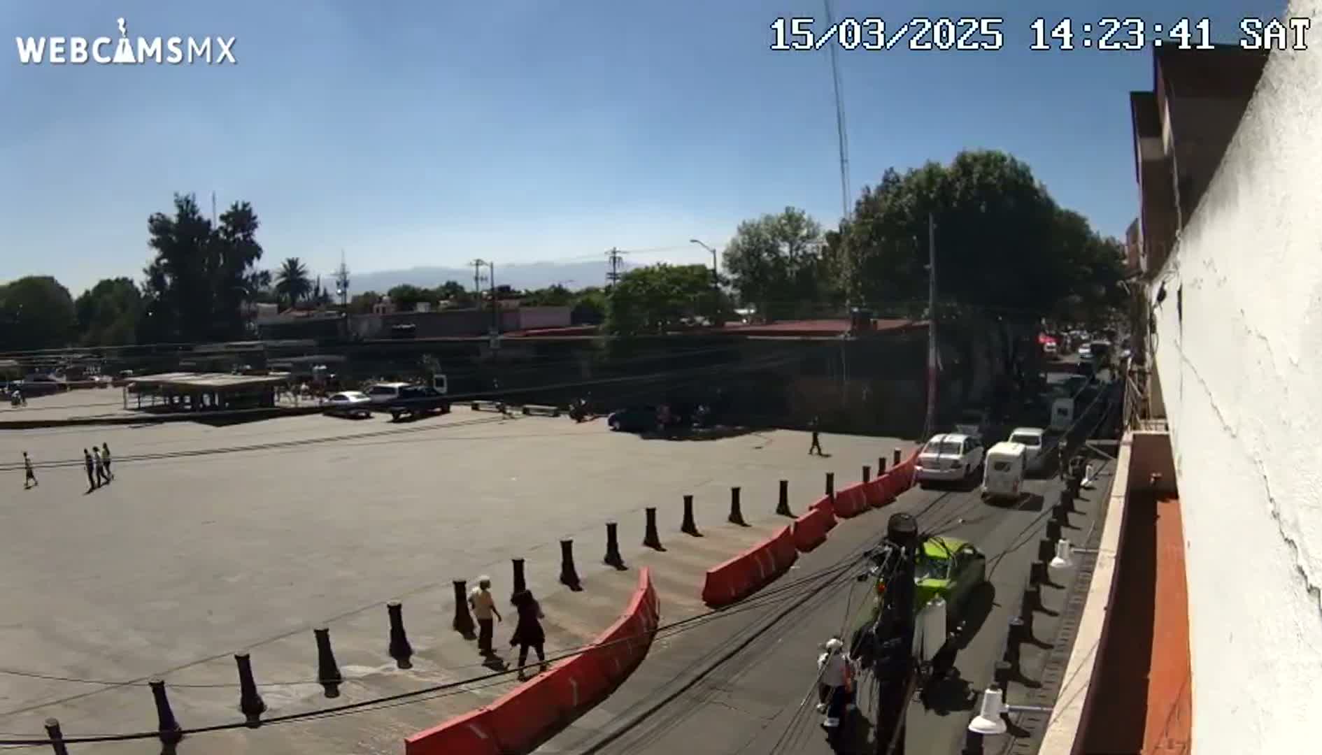 A sunny, daytime view of a large paved area with several cars and pedestrians, bordered by buildings, trees, and orange and black barriers.