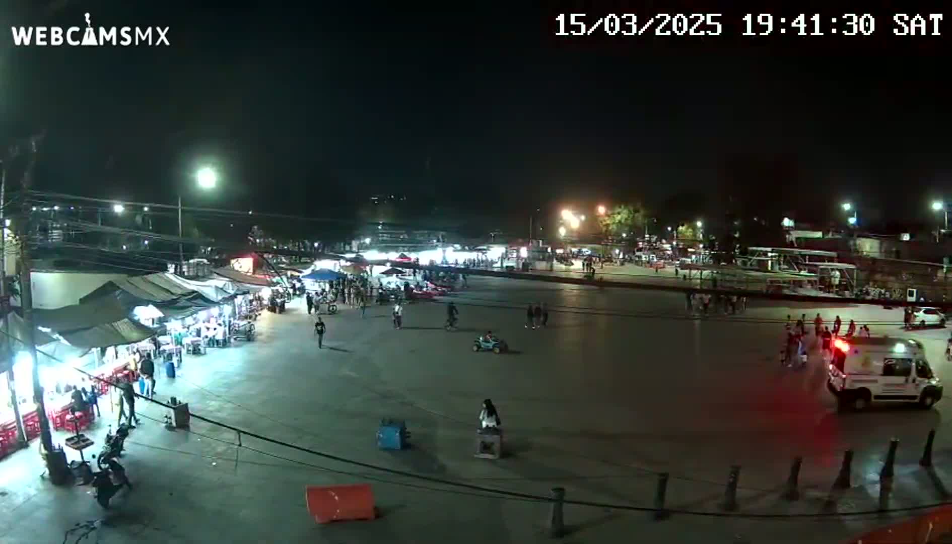 A nighttime outdoor scene shows a large plaza with numerous food stalls and people walking around, and an ambulance is present near a group of people.