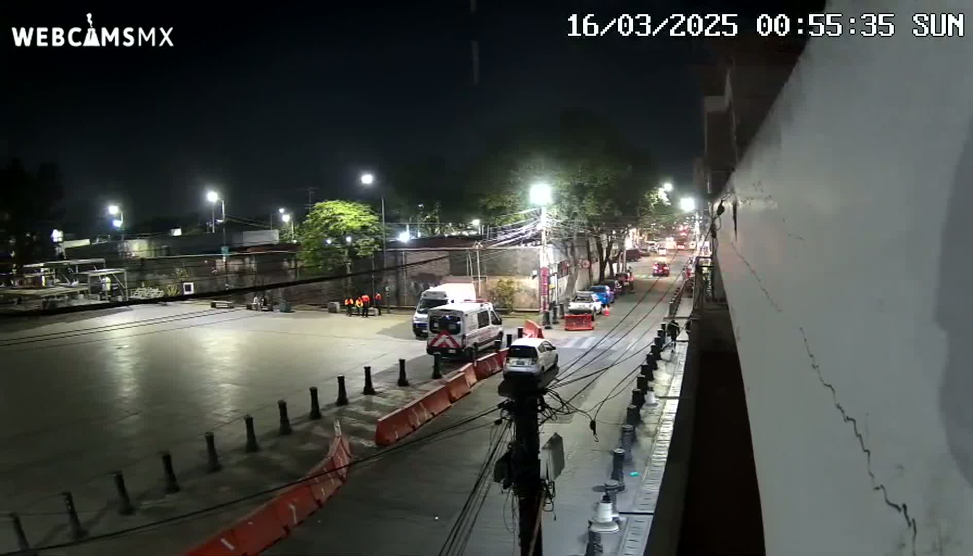 A nighttime street scene shows several parked cars, a van, and an ambulance near a roadway blocked by orange barriers, all under streetlights.