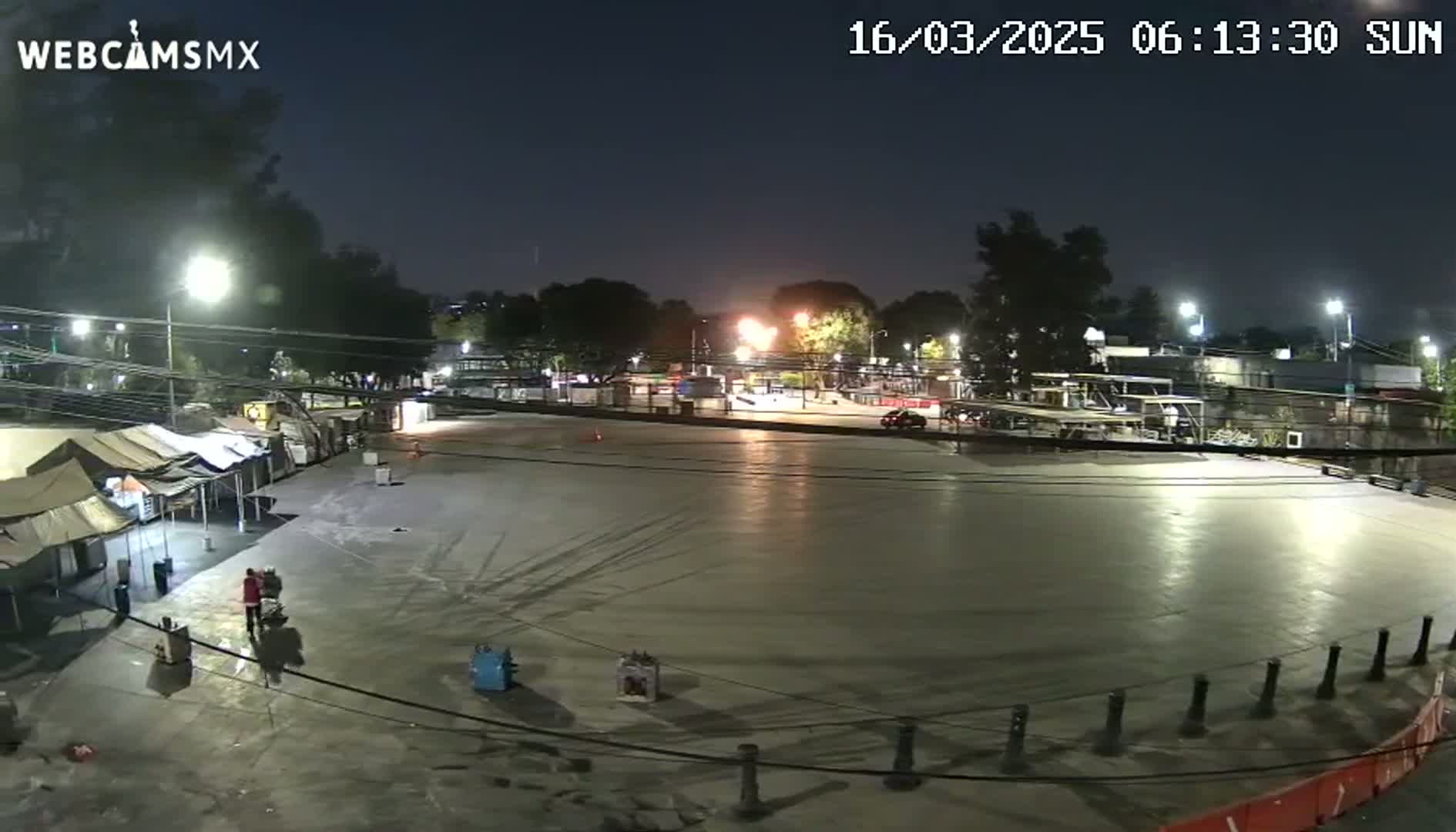 A large, empty paved area at night, illuminated by artificial lights, is surrounded by trees, buildings, and a few temporary tents, with a clear night sky.