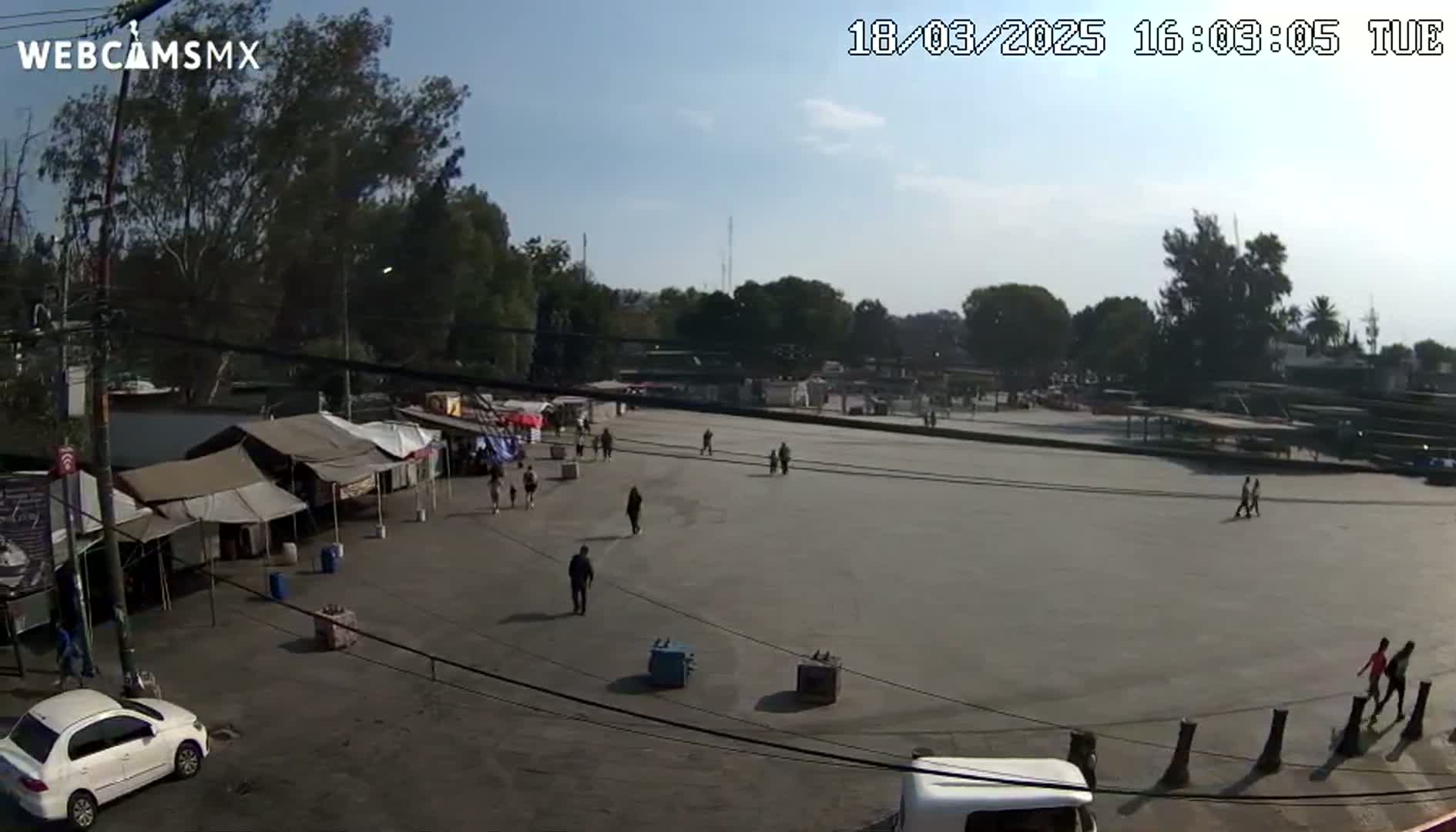 A large, paved area with several people walking around, surrounded by trees and temporary market stalls under a clear, sunny sky.