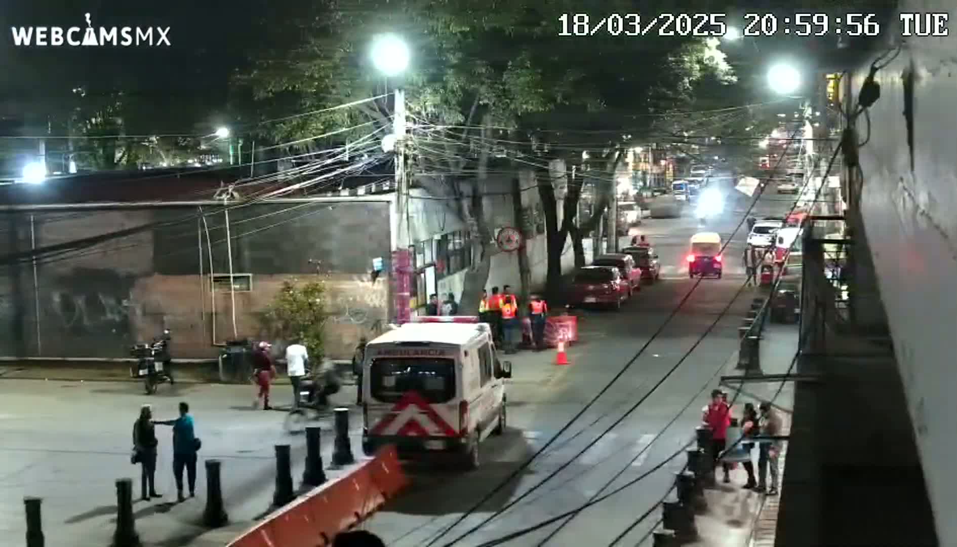 A nighttime street scene shows an ambulance and several people gathered near it, with parked cars and pedestrians visible under streetlights.
