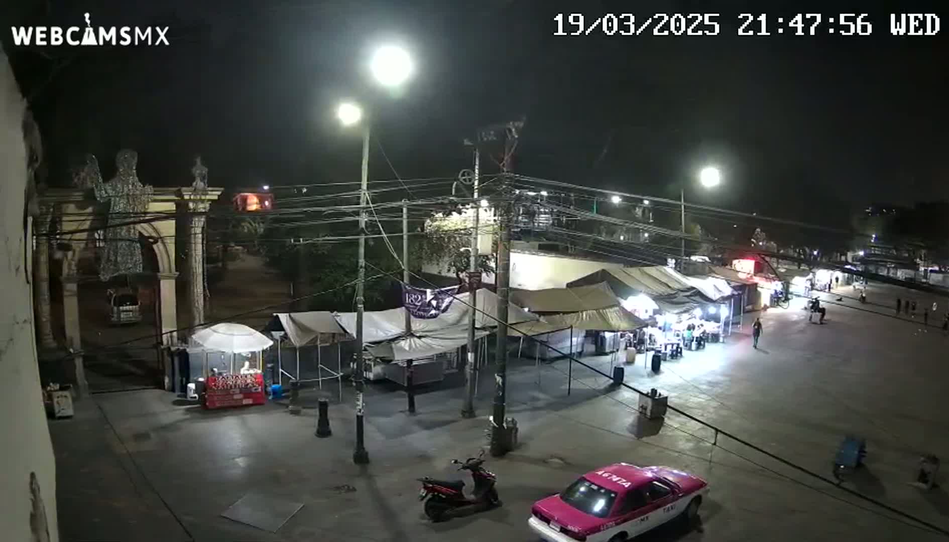 A nighttime, outdoor scene shows a plaza with several market stalls, a pink taxi, and a scooter, all under bright lights.