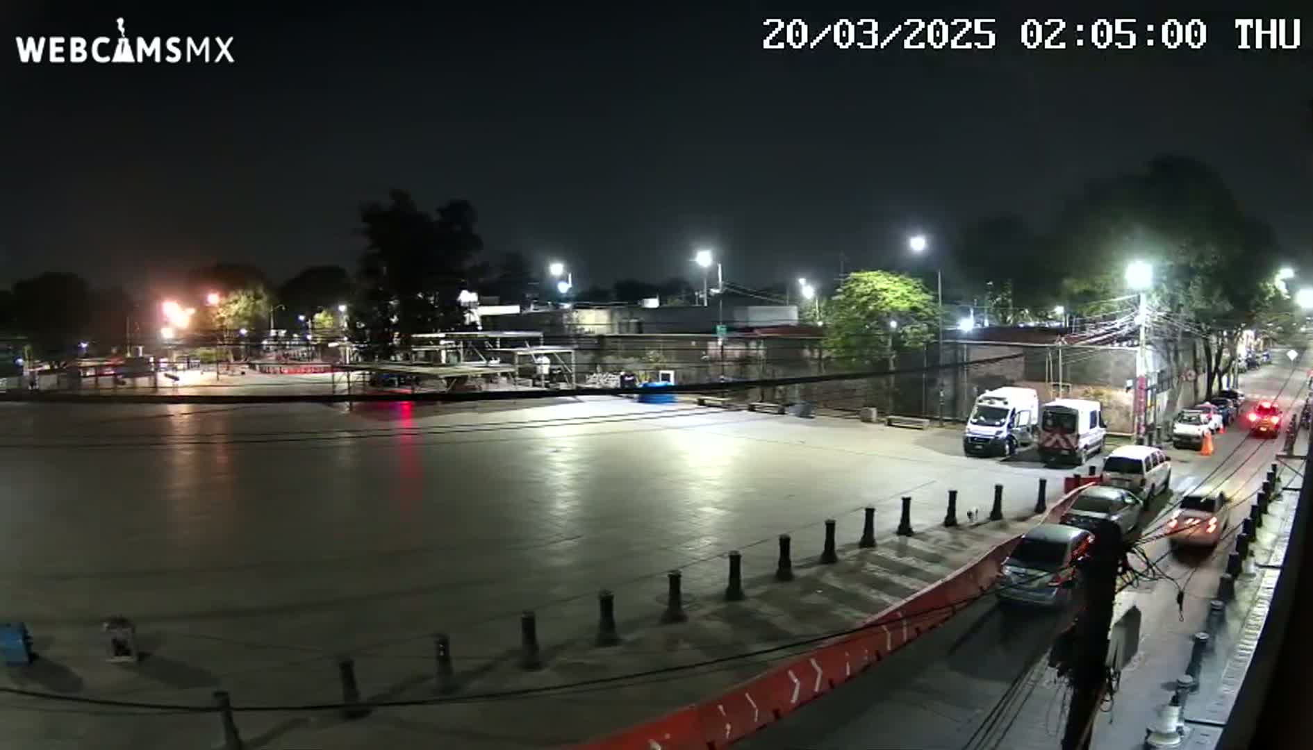 A nighttime view of an empty plaza with several cars parked and driving on a nearby street, illuminated by streetlights.