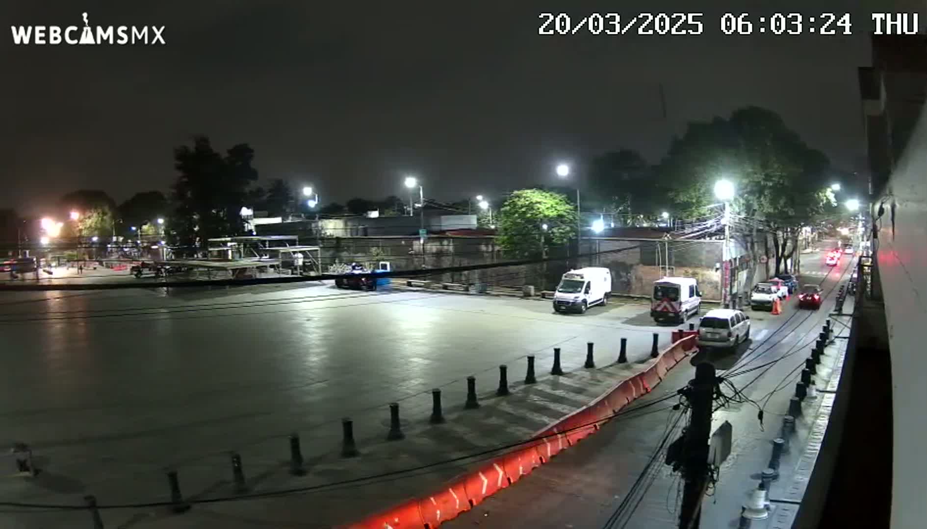 A nighttime view of a mostly empty paved lot with several vehicles, including vans and cars, parked and driving on adjacent streets, under illuminated streetlights.
