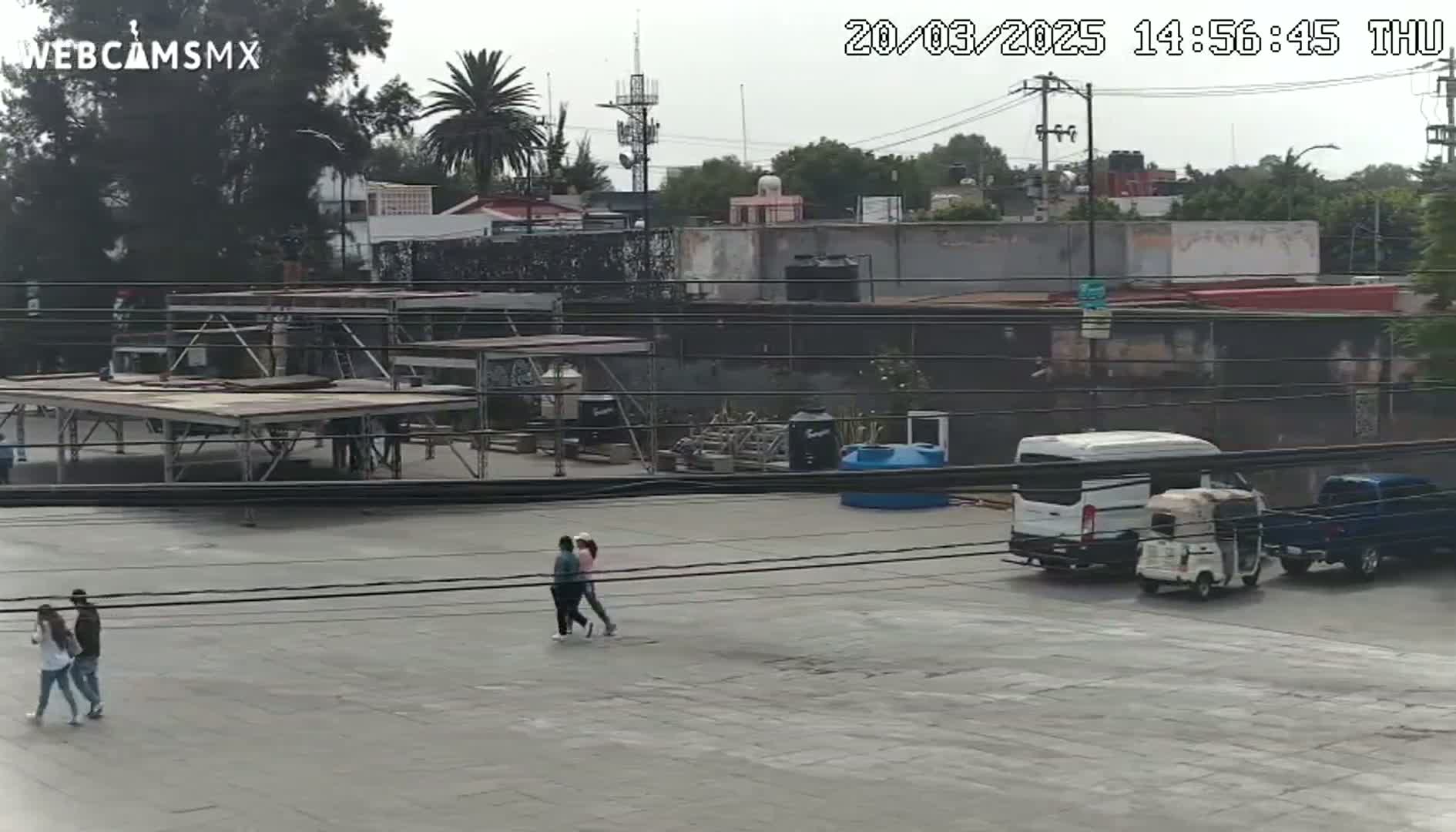 An overcast day reveals a paved area with several people walking, a white van, a three-wheeled vehicle, and a blue pickup truck parked near some buildings and temporary staging structures.