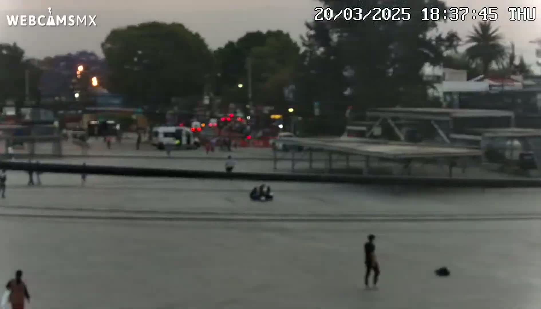 A mostly empty plaza with a few pedestrians and a group of people sitting on the ground, under an overcast sky at dusk.
