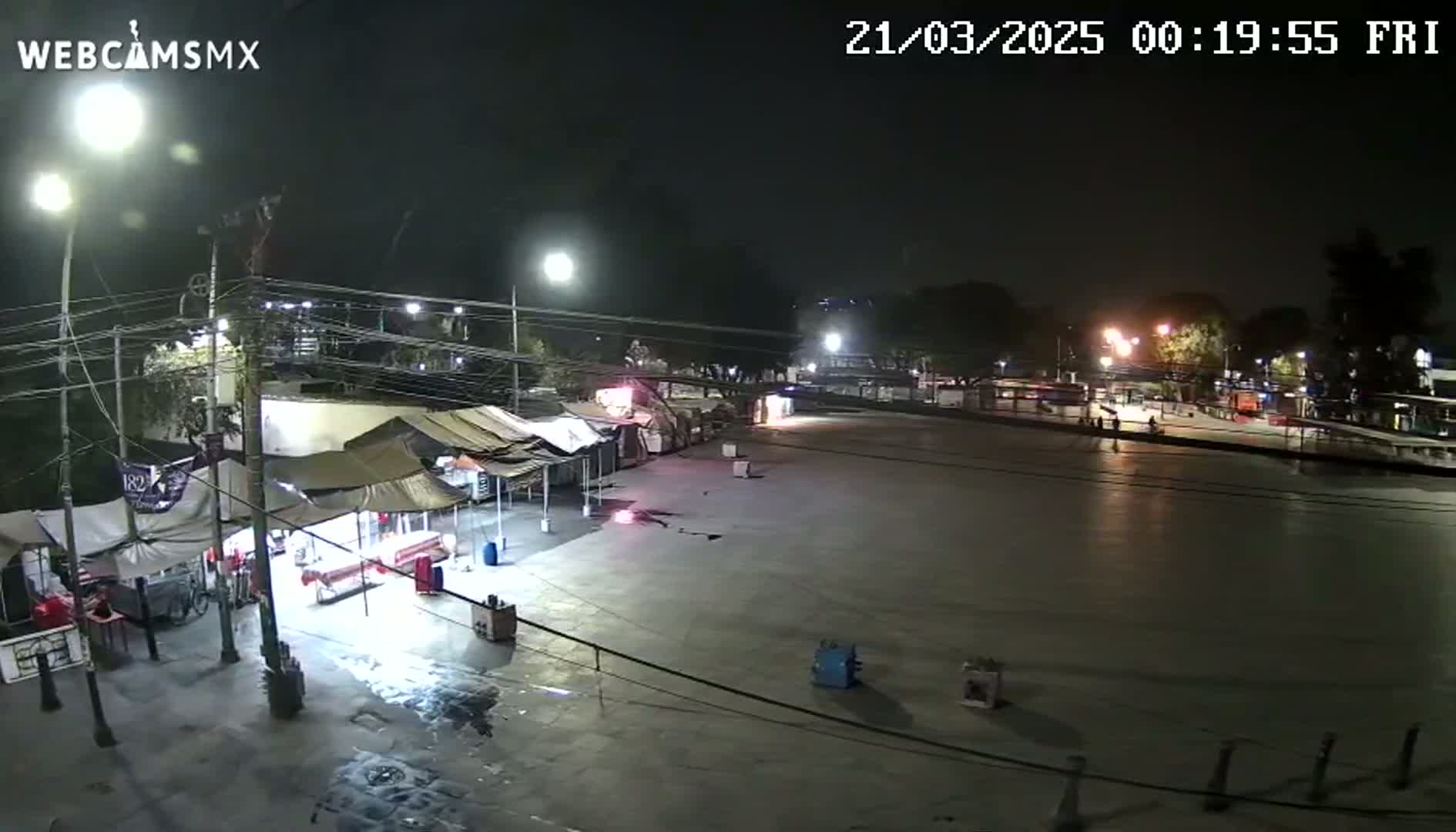 A nighttime view of a plaza with several food stalls and some people, under clear skies.