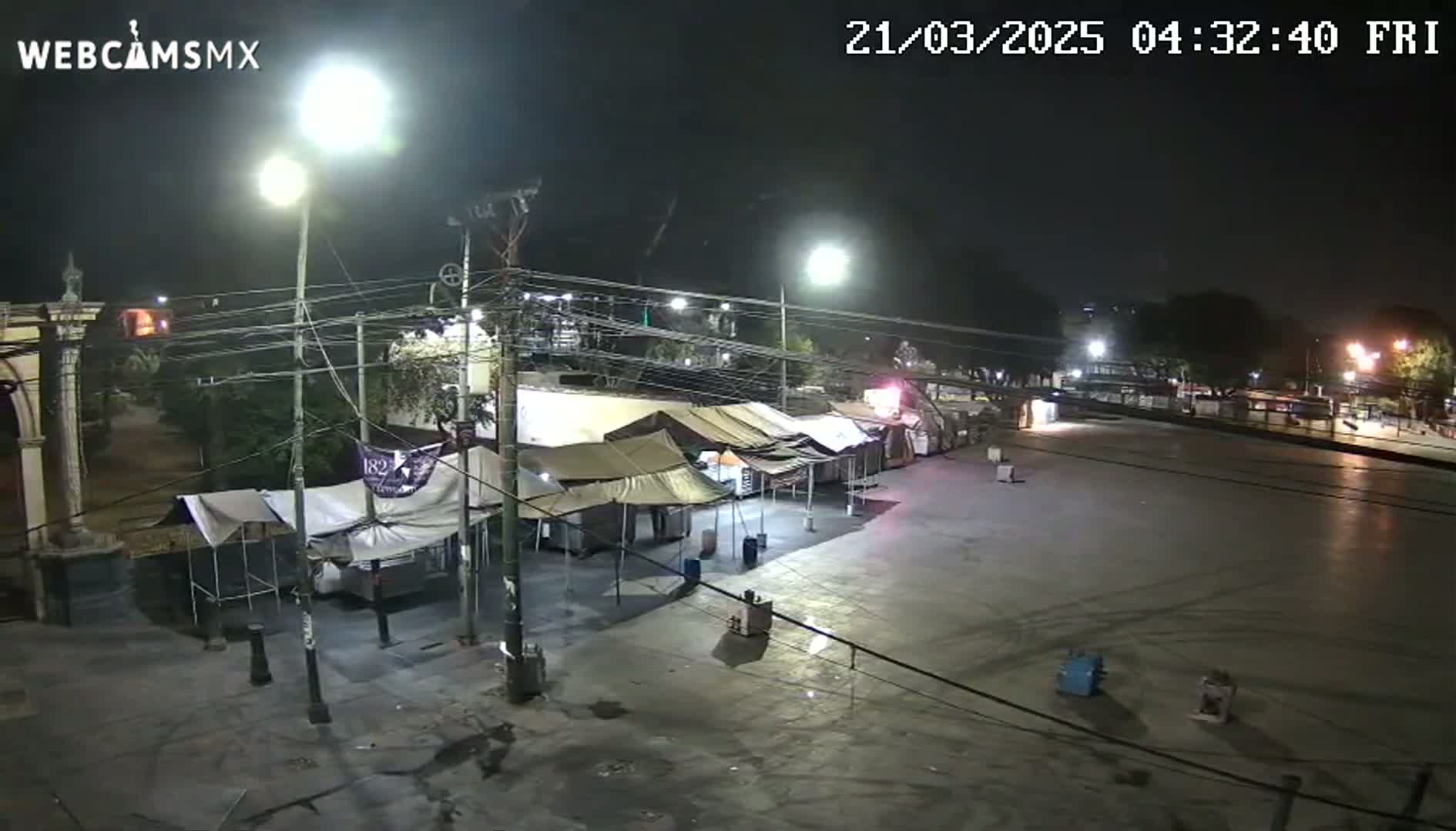 A nighttime view of an empty plaza with several tents set up, illuminated by streetlights under a dark sky.