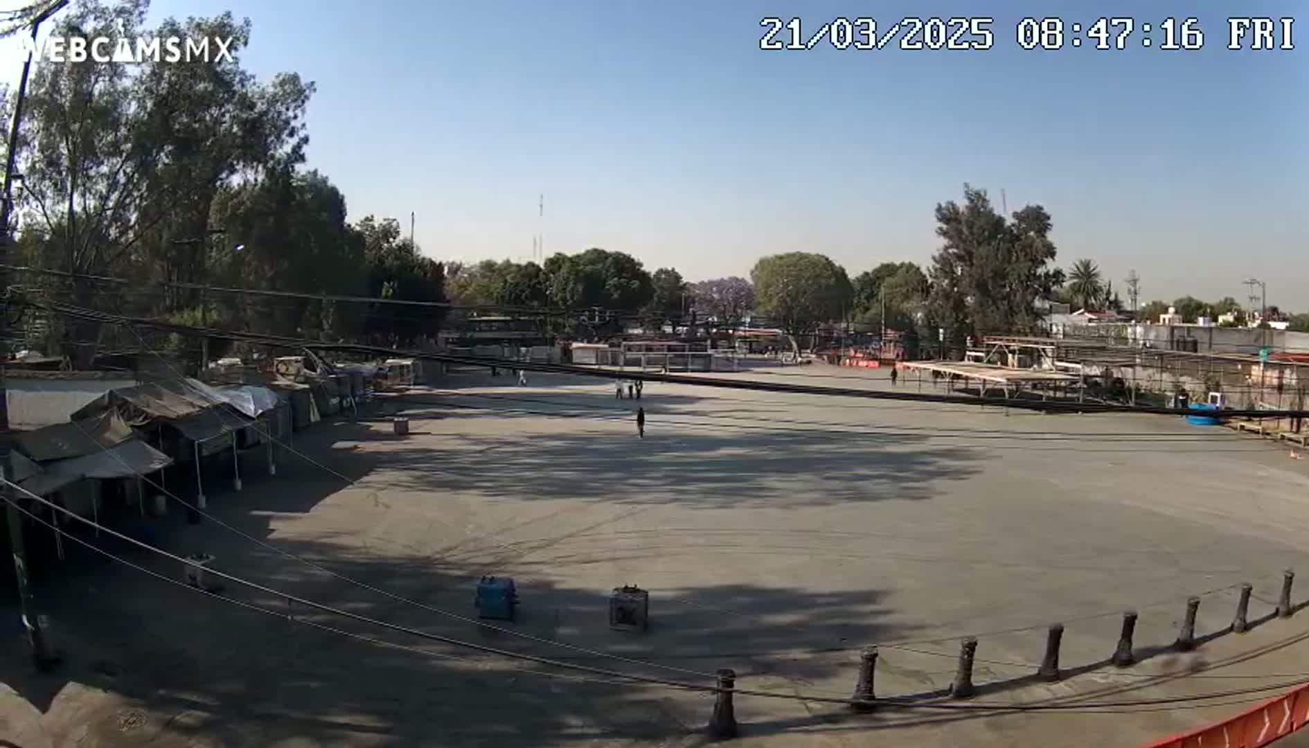 A large, empty paved area, surrounded by trees and temporary structures, is seen under a clear, sunny sky.
