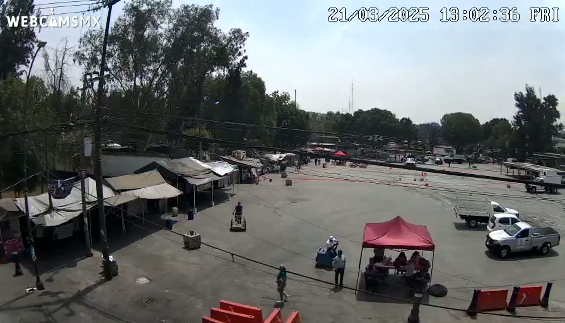 A mostly empty paved area with several market stalls and tents under a partly cloudy sky, surrounded by trees and parked vehicles.