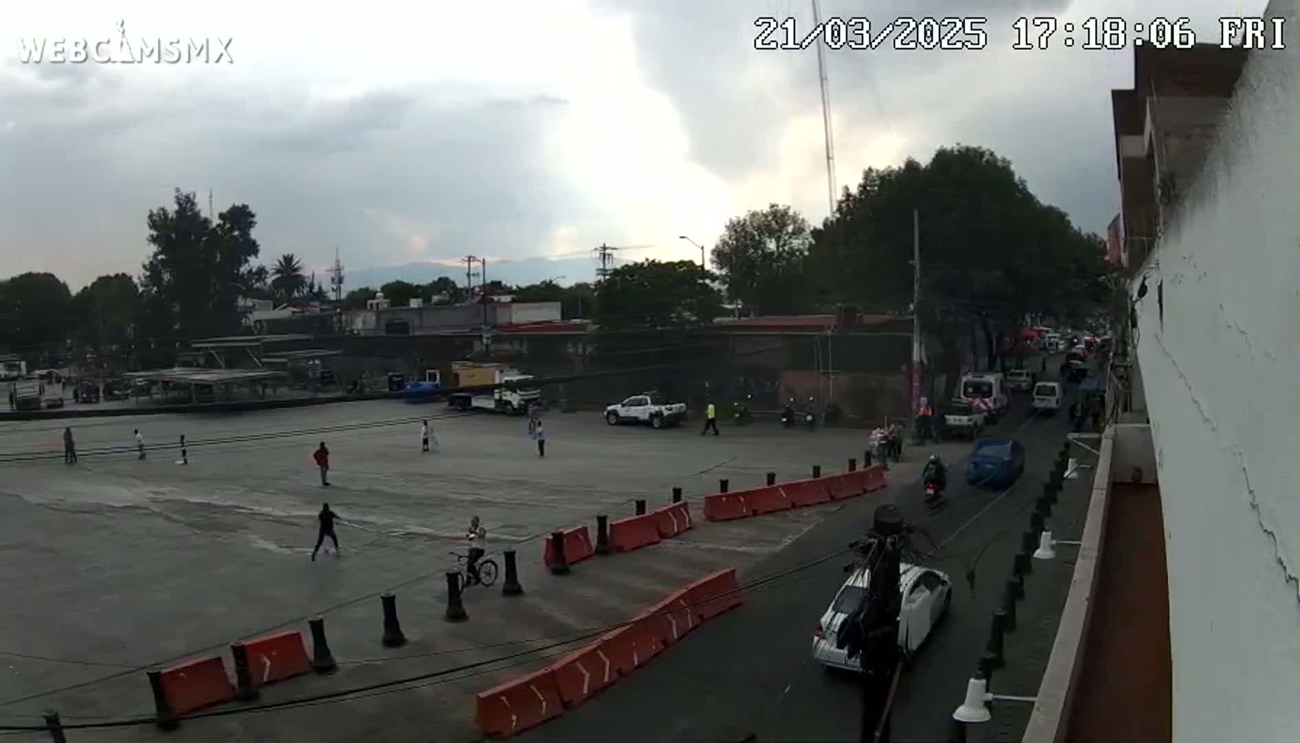 A mostly empty plaza with several people scattered about and vehicles driving on a street next to a building under an overcast sky.