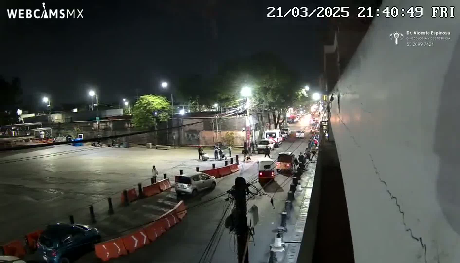 A nighttime view of a street scene with several cars and people, illuminated by streetlights.