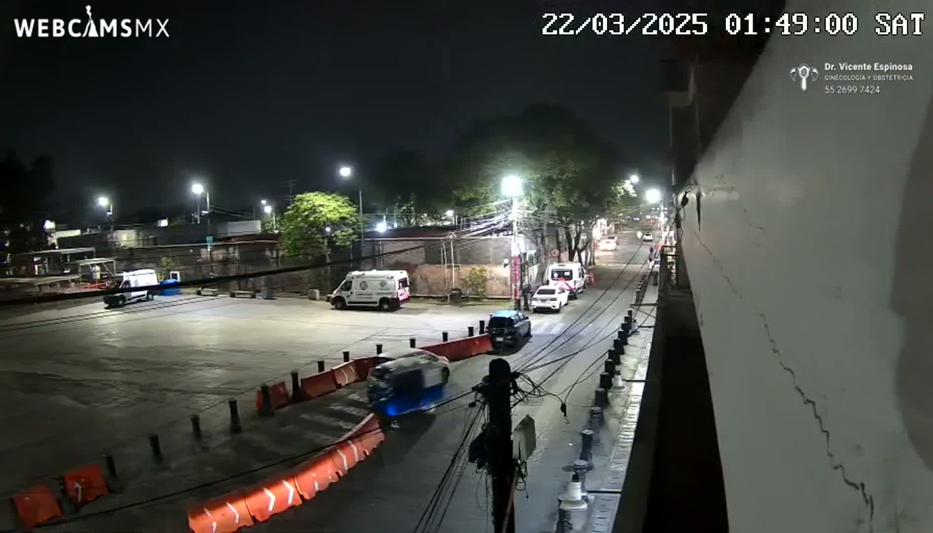 A nighttime street scene shows several vehicles parked and driving on a road lined with streetlights and orange barriers, under clear skies.