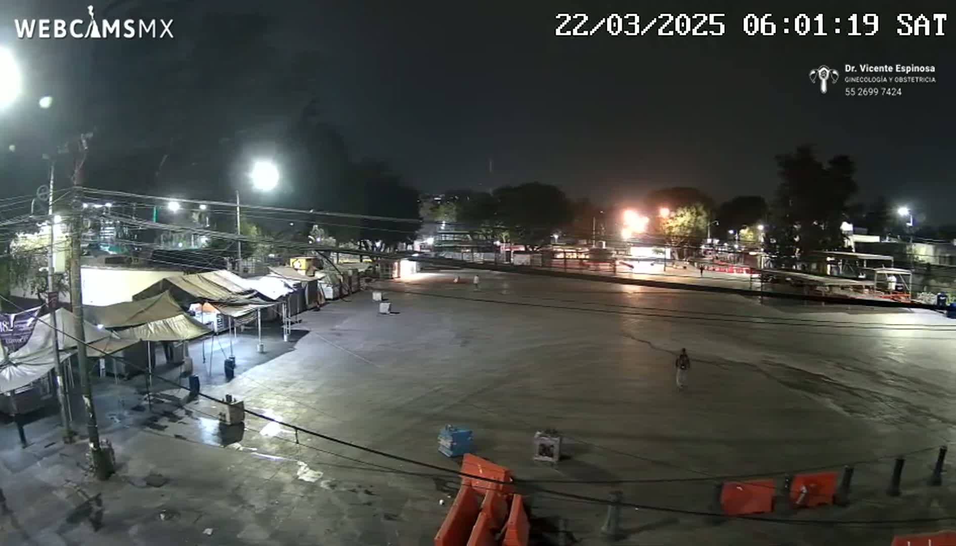 A nighttime view of a mostly empty plaza with several market stalls set up along one side, under partly cloudy skies.