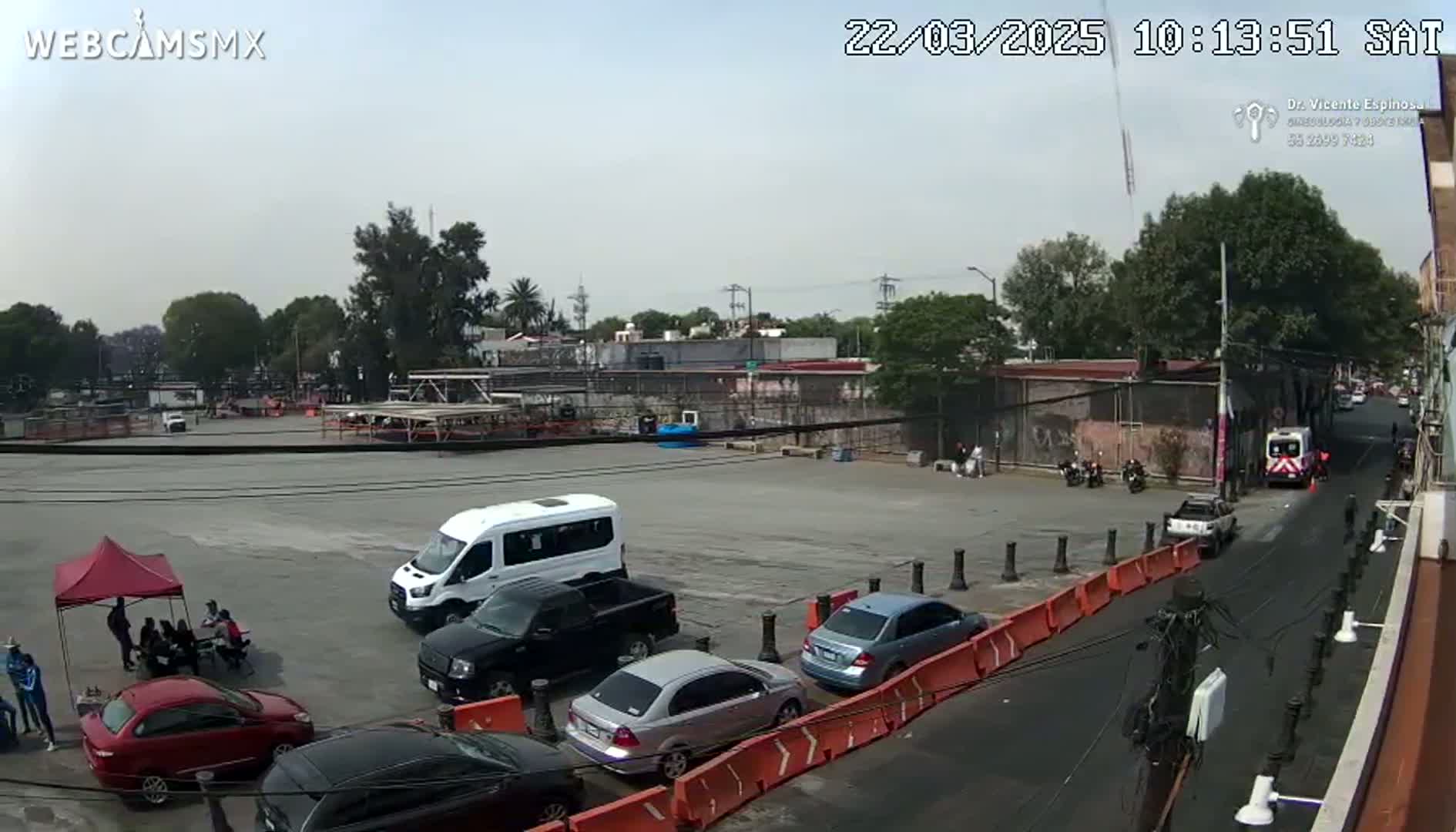 A mostly empty paved lot with several cars and a van parked, a red tent with people sitting underneath, and a few motorcycles parked nearby, under a mostly clear sky.