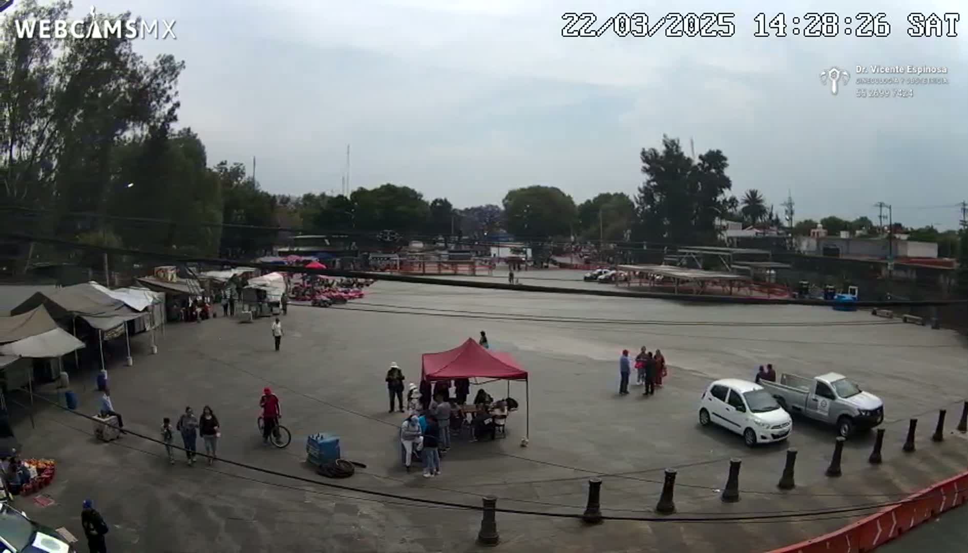 An overcast day reveals a large plaza with several small tents, food vendors, people walking around, and a few vehicles parked, all surrounded by trees and buildings.