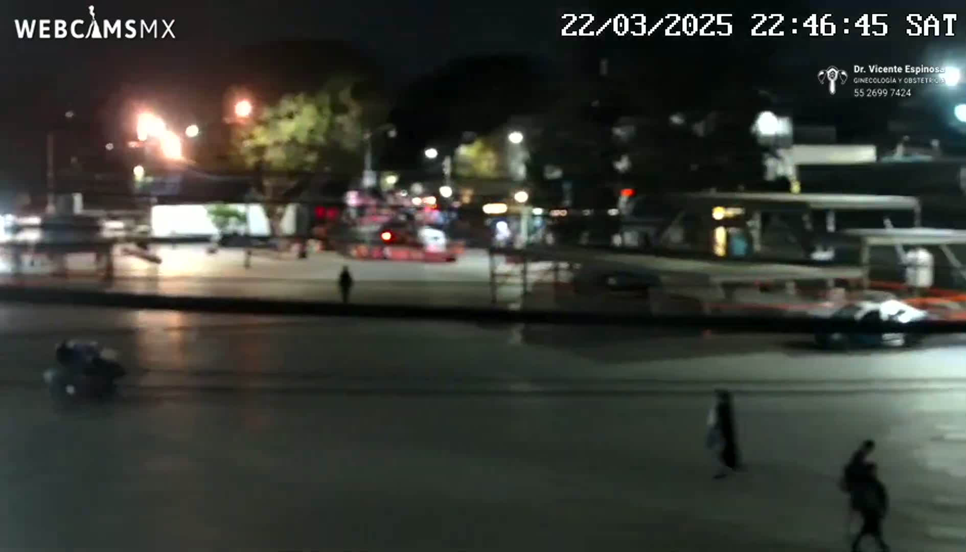 A nighttime, outdoor view of a mostly empty plaza with a few pedestrians and vehicles, under clear skies.