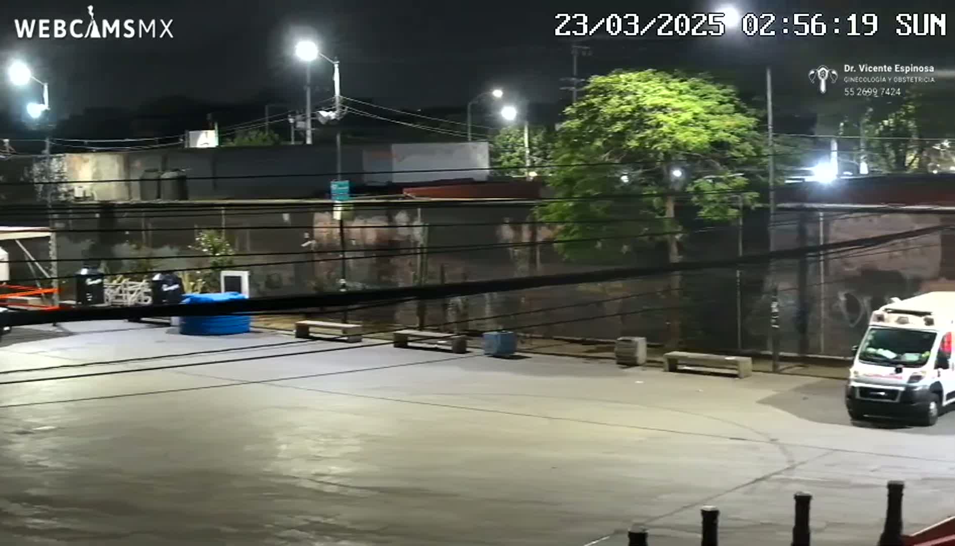 A nighttime view of an empty paved area, illuminated by streetlights, with a white van parked near a fence and several objects scattered about.