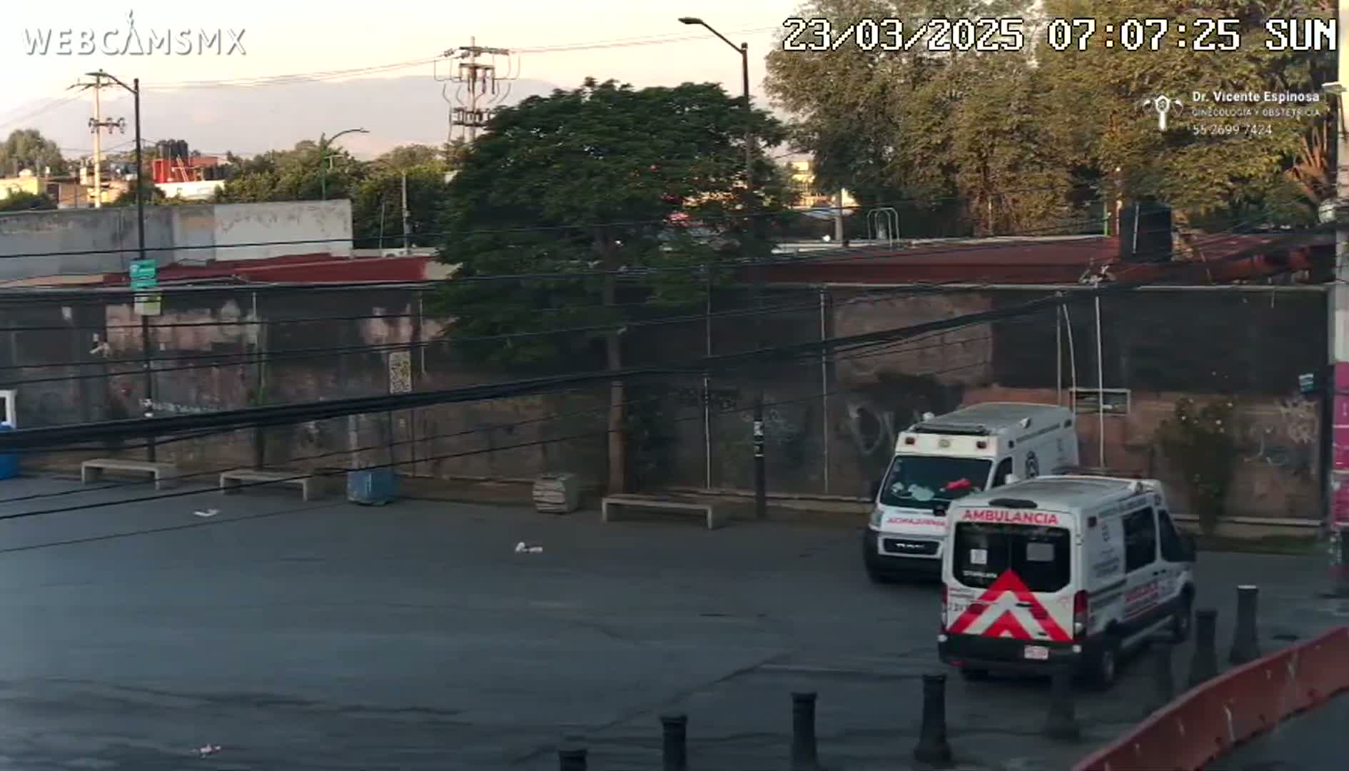 Two ambulances are parked in a paved lot next to a graffiti-covered wall under a partly cloudy sky at sunrise.