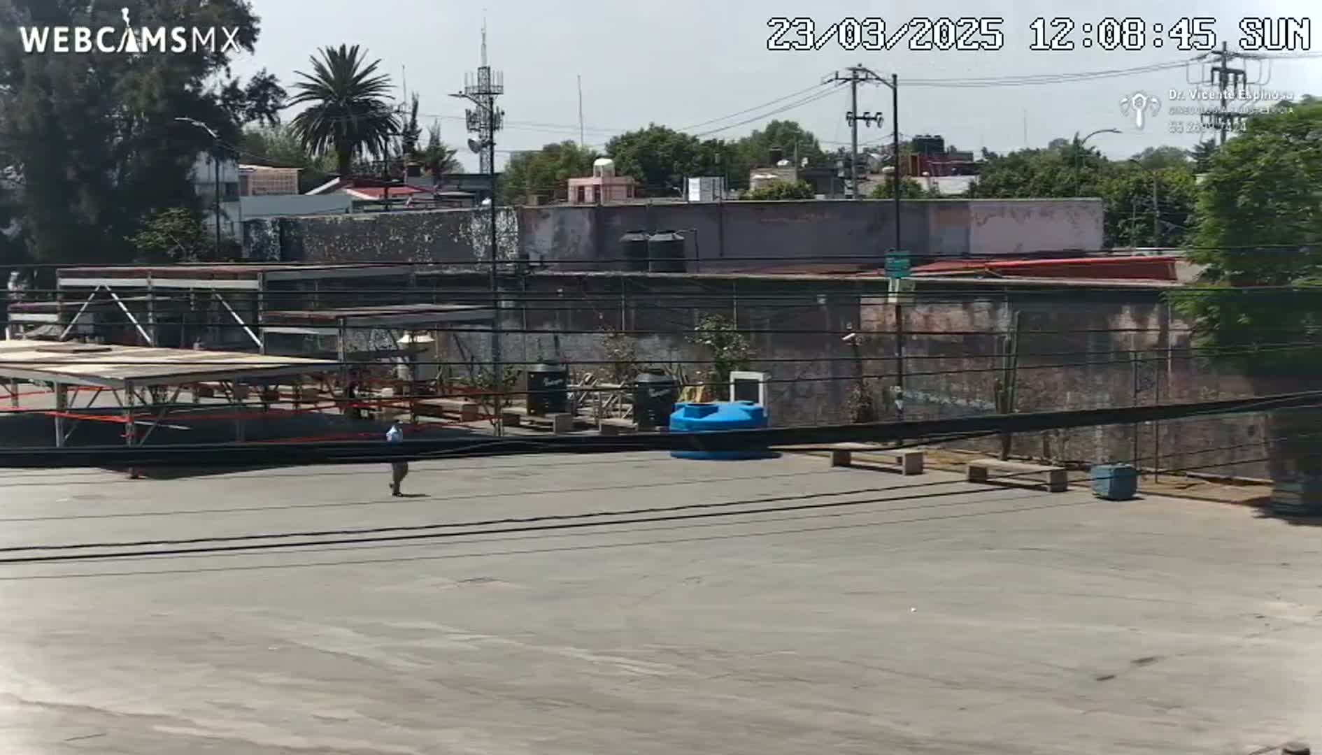 A person walks across a paved area in front of buildings under a sunny sky.