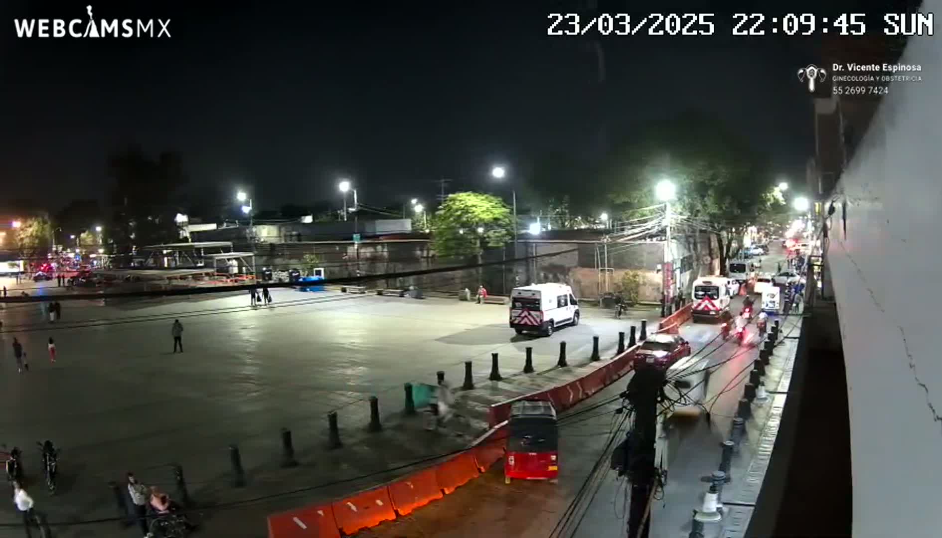 A nighttime, city street scene shows a mostly empty plaza with a few pedestrians, several vehicles including ambulances, and a red auto rickshaw near orange barriers.