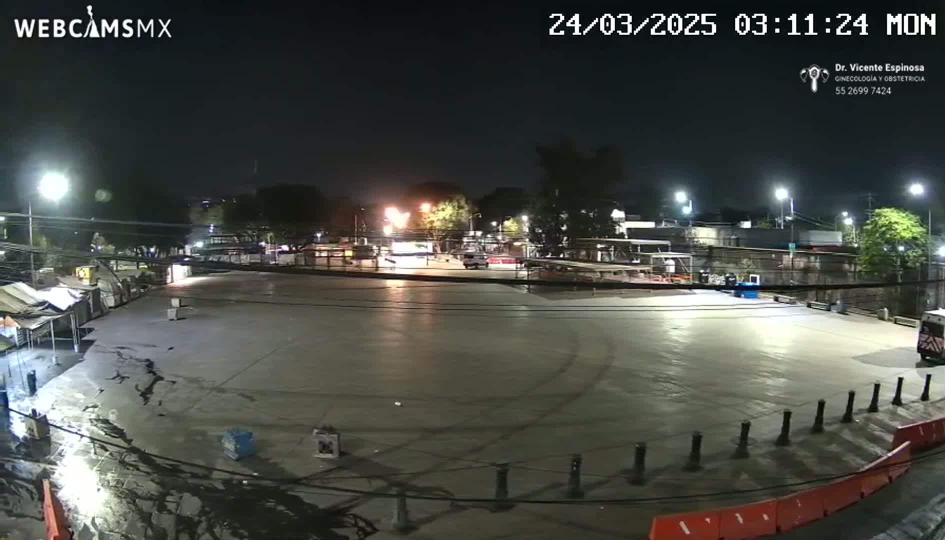 A large, empty paved lot at night is illuminated by streetlights, with a few structures and vehicles visible in the distance.