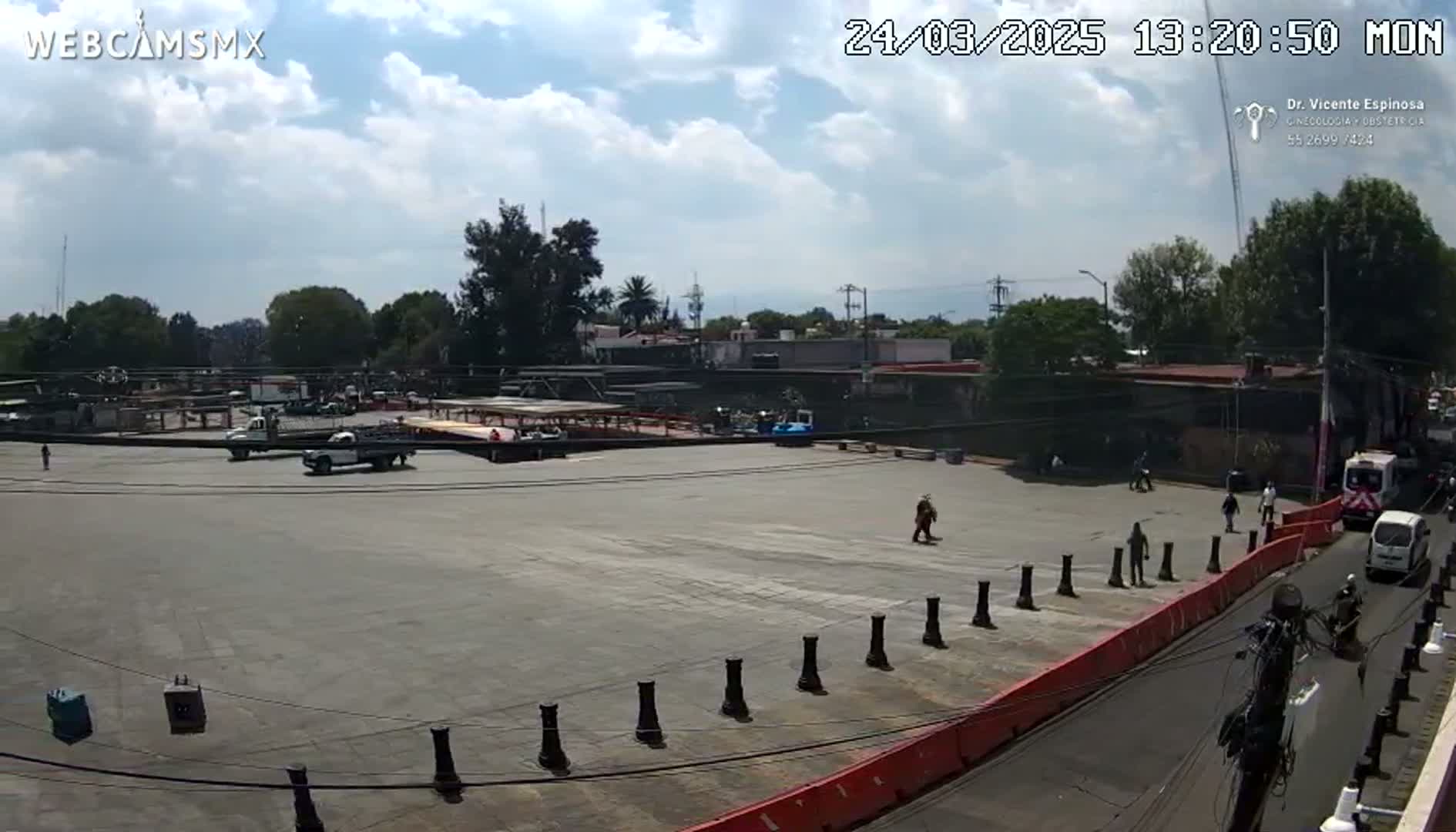 A mostly empty paved area with a few vehicles and pedestrians, under a partly cloudy sky.