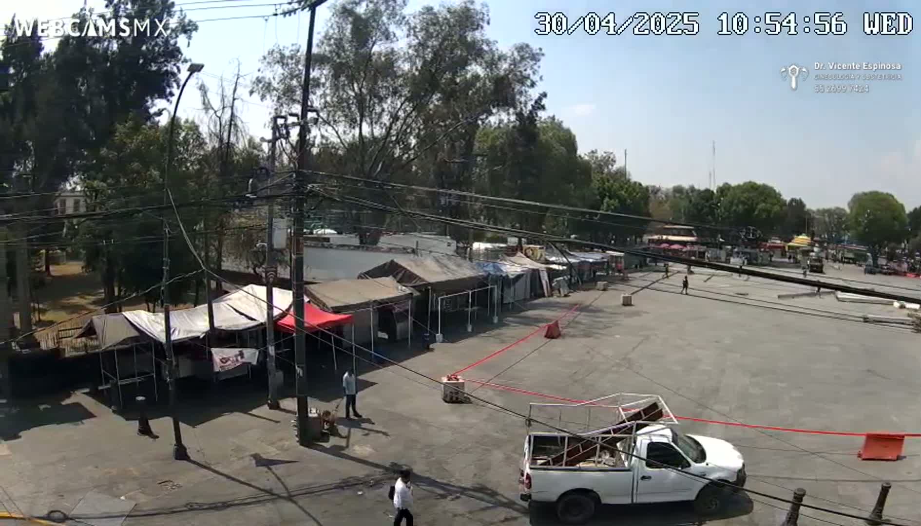 A mostly sunny outdoor scene shows a paved area with numerous market stalls and tents, a few people walking around, and a white pickup truck parked nearby.