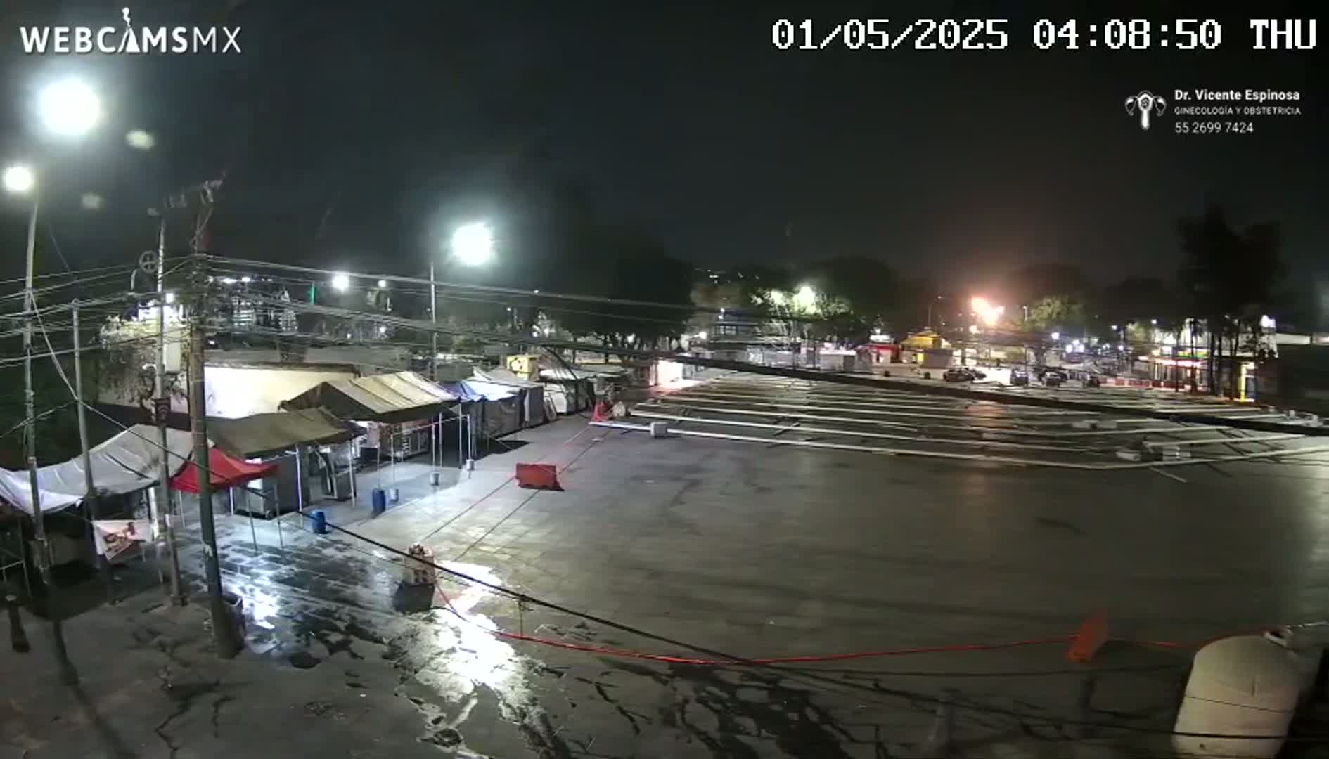 A nighttime view of a wet, empty parking lot surrounded by tents and buildings under streetlights.