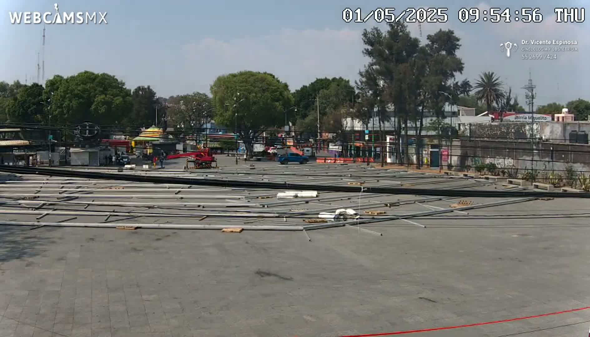 Under a clear sky, a large paved area is scattered with long metal beams and building materials near several buildings and trees.