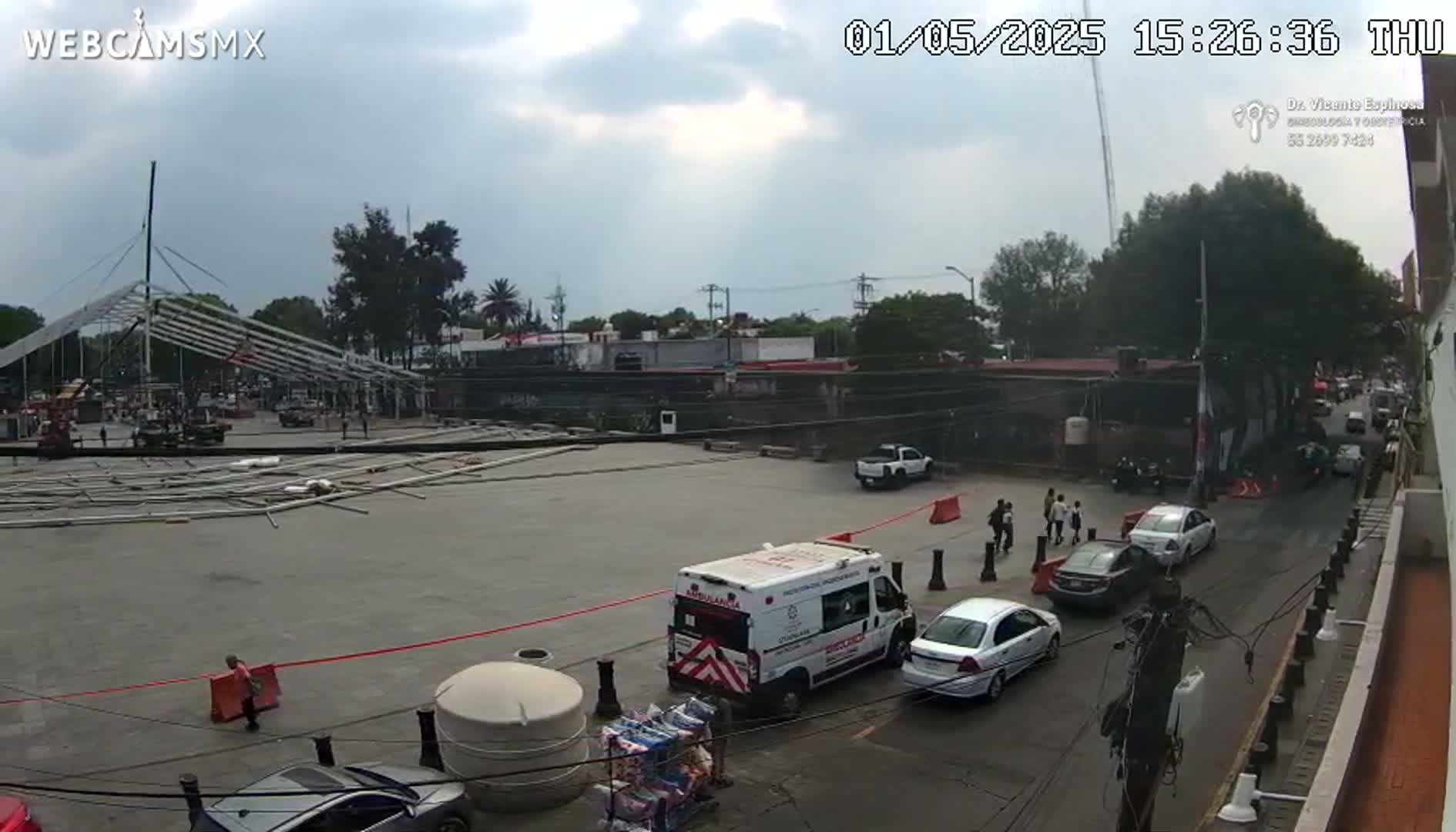 A mostly cloudy day reveals an outdoor scene of a large, empty paved area with disassembled metal scaffolding, a few vehicles including an ambulance, and several people standing near a street with cars parked along the side.