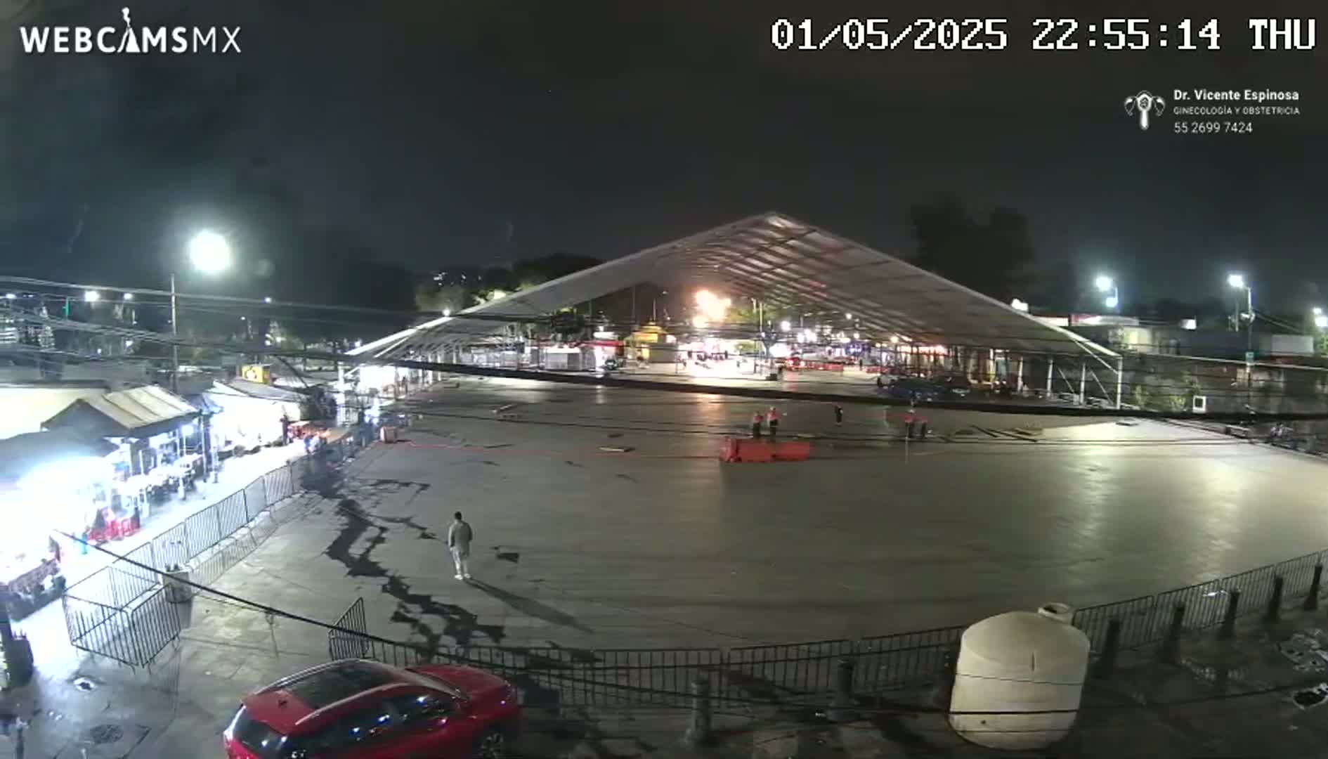 A nighttime, outdoor view of a large, empty paved area under a temporary tent structure, surrounded by smaller buildings and fences, with a few people visible.