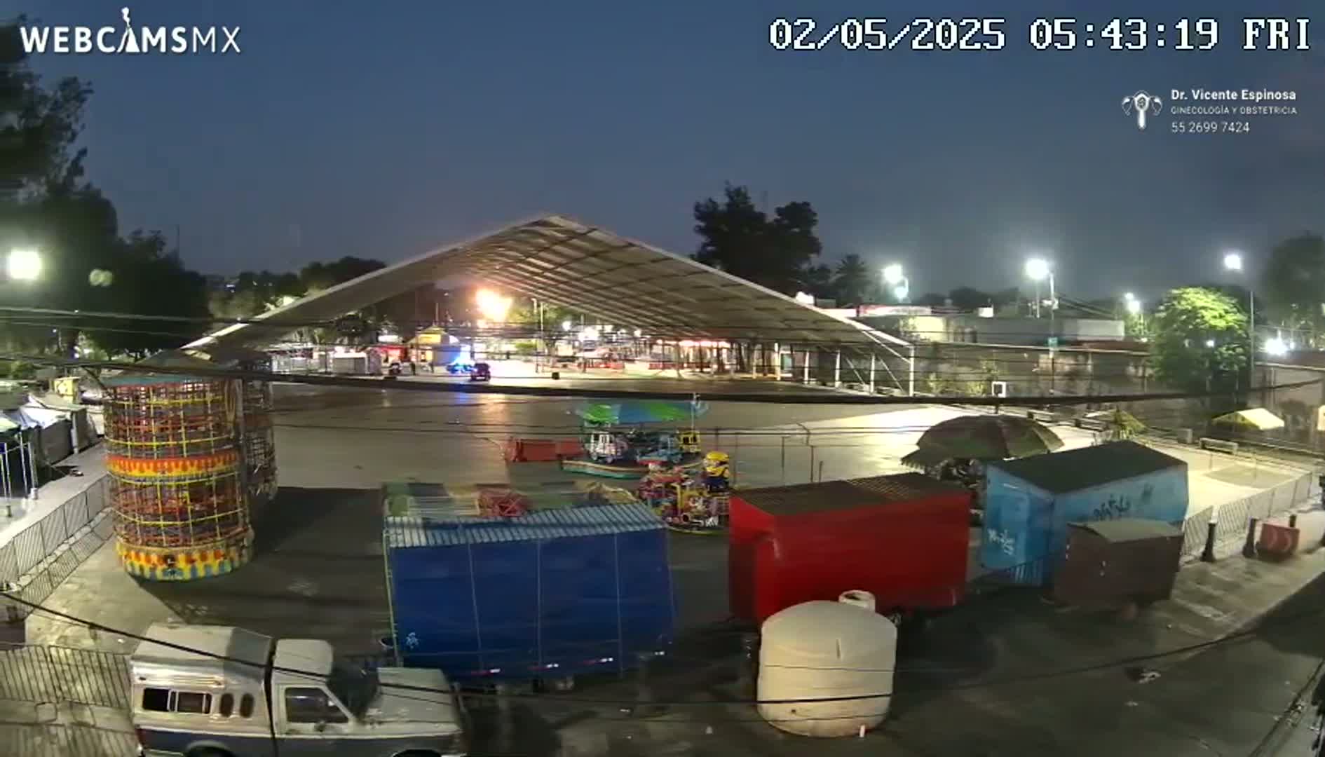 An outdoor nighttime scene shows a paved area with carnival rides and food stalls under a large metal awning, illuminated by artificial lights.