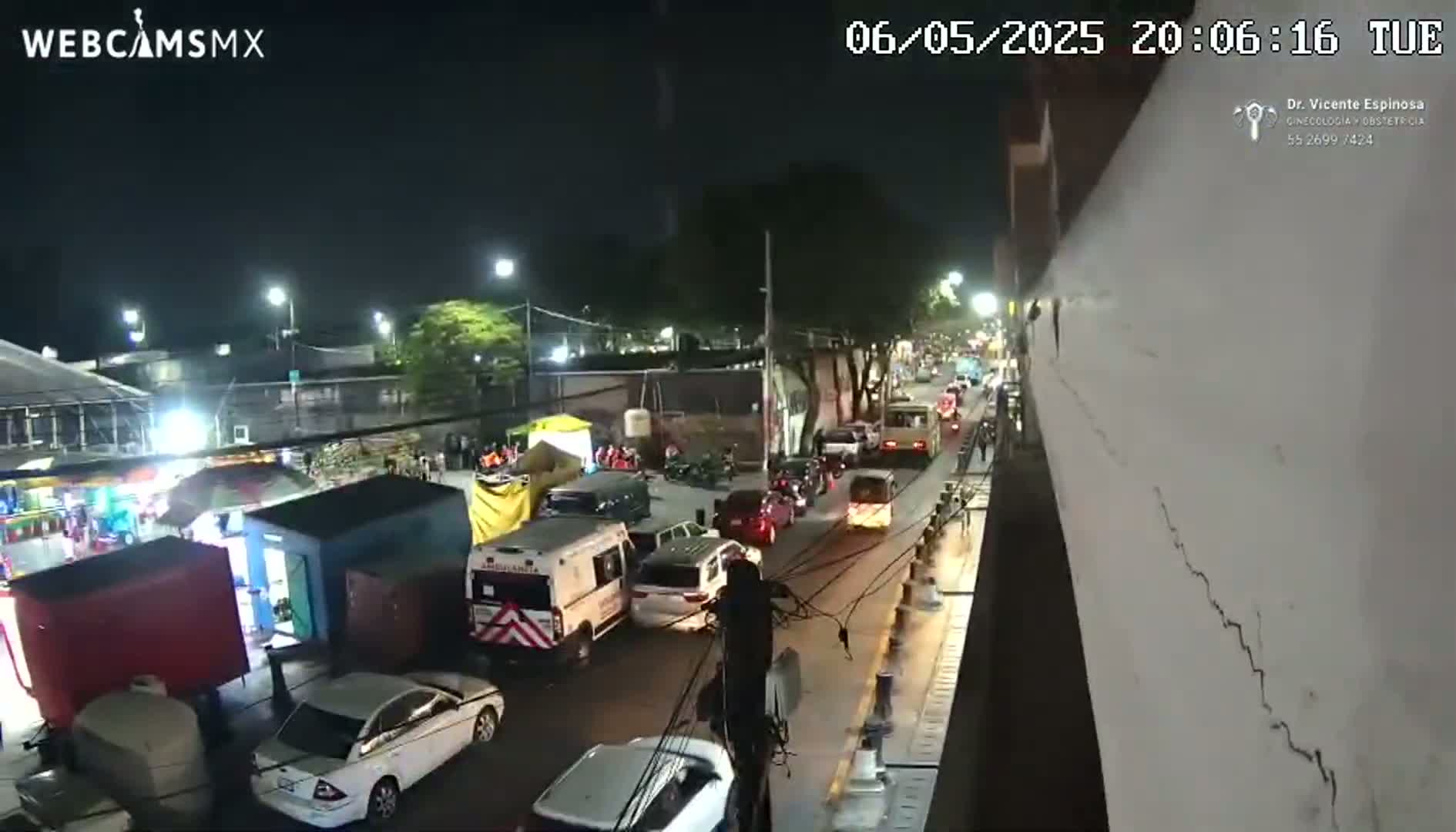 A nighttime, street-level view shows a street with cars and an ambulance, next to a fair or market with several stalls and rides under streetlights.