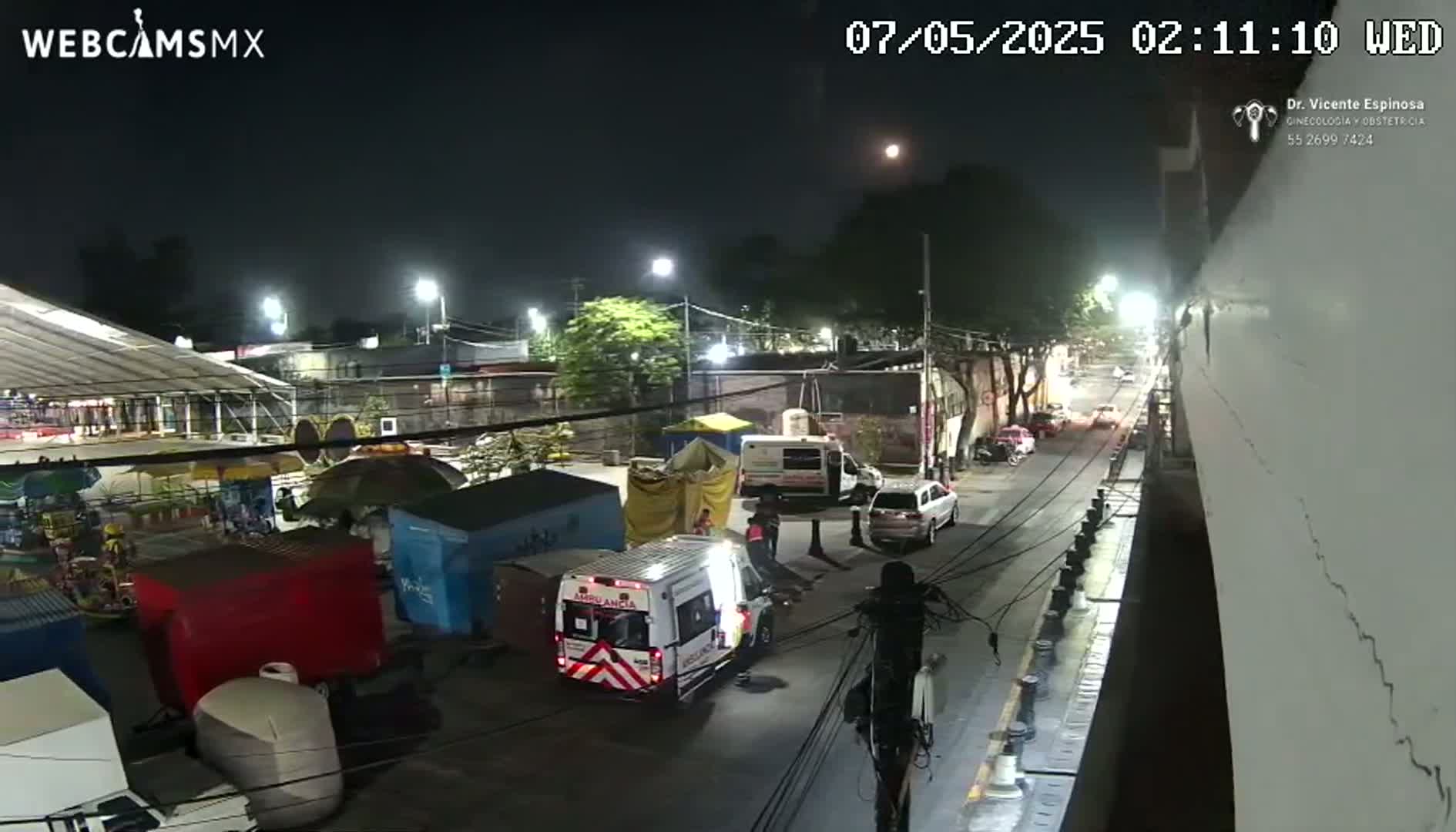 A nighttime street scene shows an ambulance, other vehicles, and several food stalls and carnival-like structures, under a clear sky with a visible moon.
