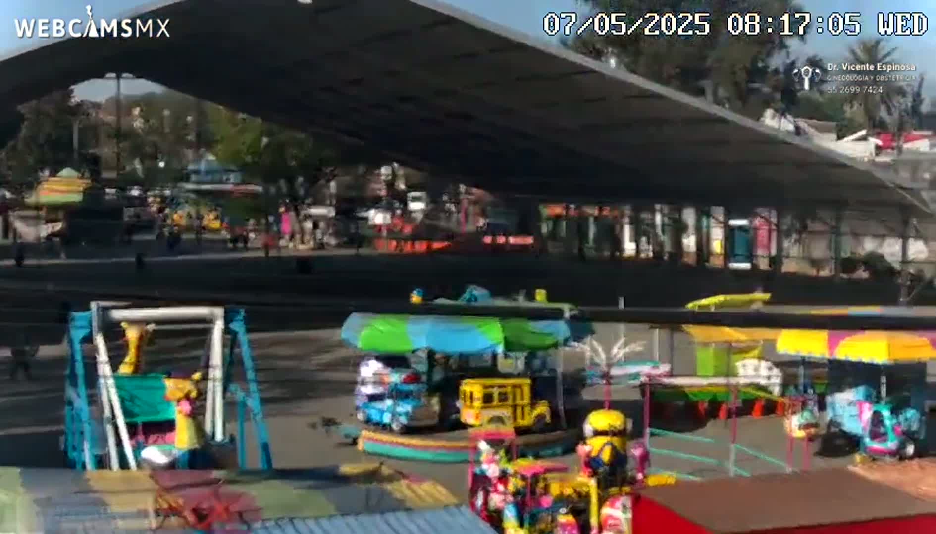 Under a large, shaded structure on a sunny day, several colorful carnival rides are visible in an open paved area with sparse crowds of people.