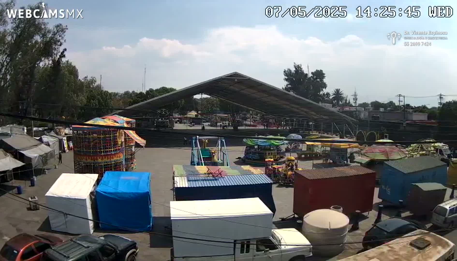 A fairground with various rides and game stalls is situated under a large metal roof outdoors on a partly cloudy day.