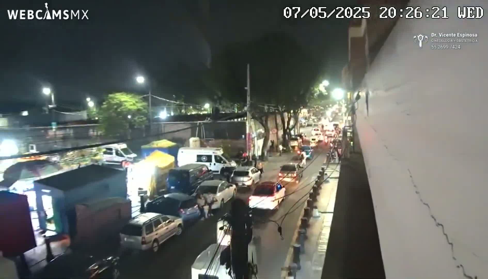 A nighttime street scene shows a line of slow-moving cars and pedestrians near several small roadside stands under streetlights.