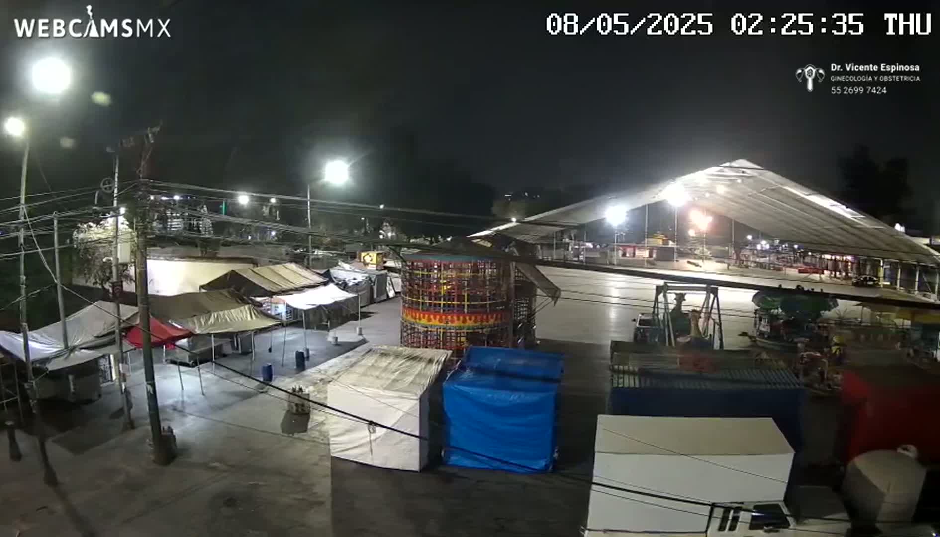 A nighttime outdoor scene shows a fairground with several tents, carnival rides, and shipping containers under a large, illuminated tent structure.