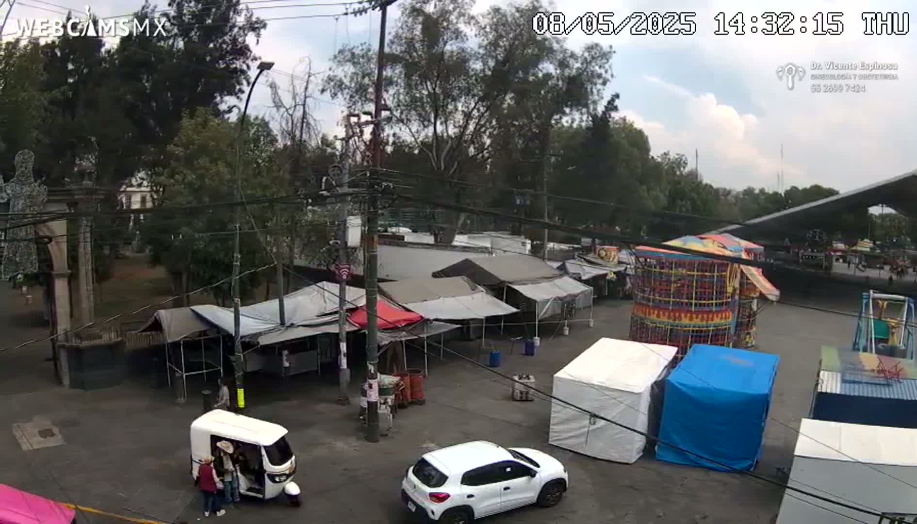 An outdoor market scene under a partly cloudy sky features numerous tents, a small amusement park ride, and several vehicles, including a white car and a three-wheeled vehicle.